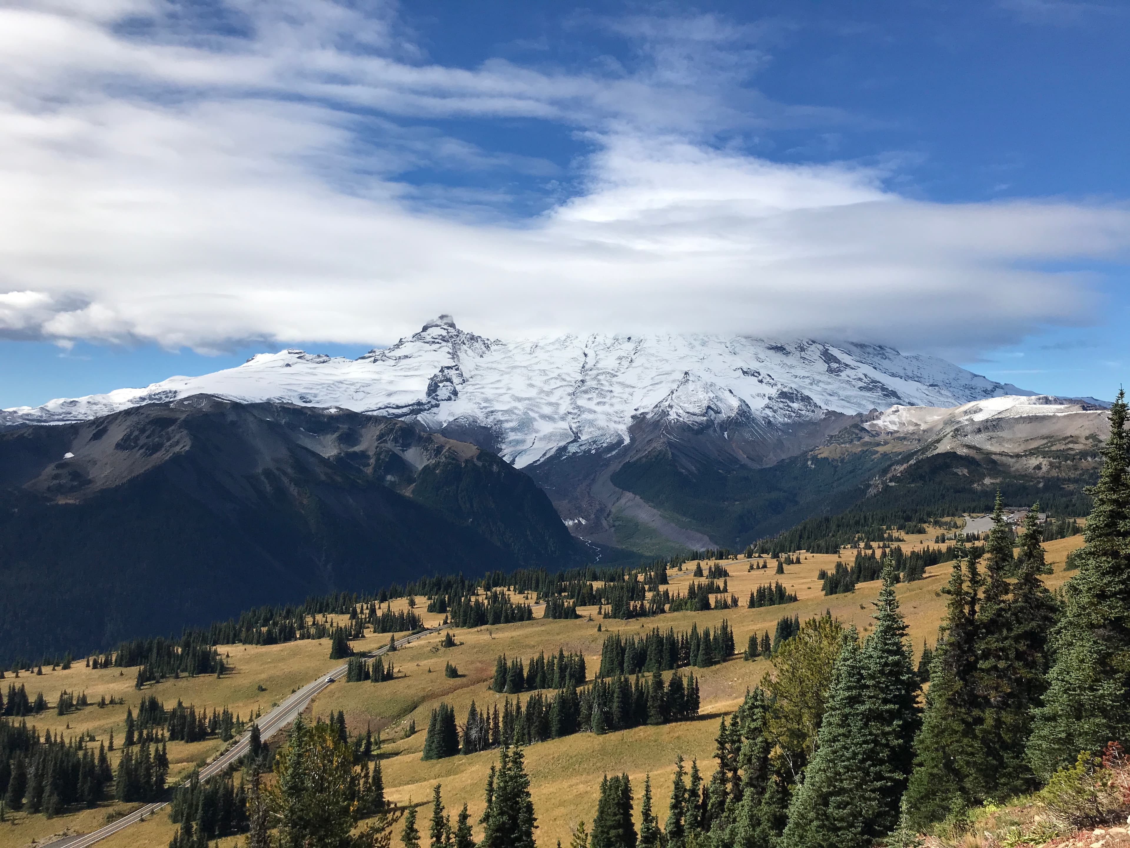 A road curves through subalpine meadows underneath a glaciated peak wrapped in clouds.