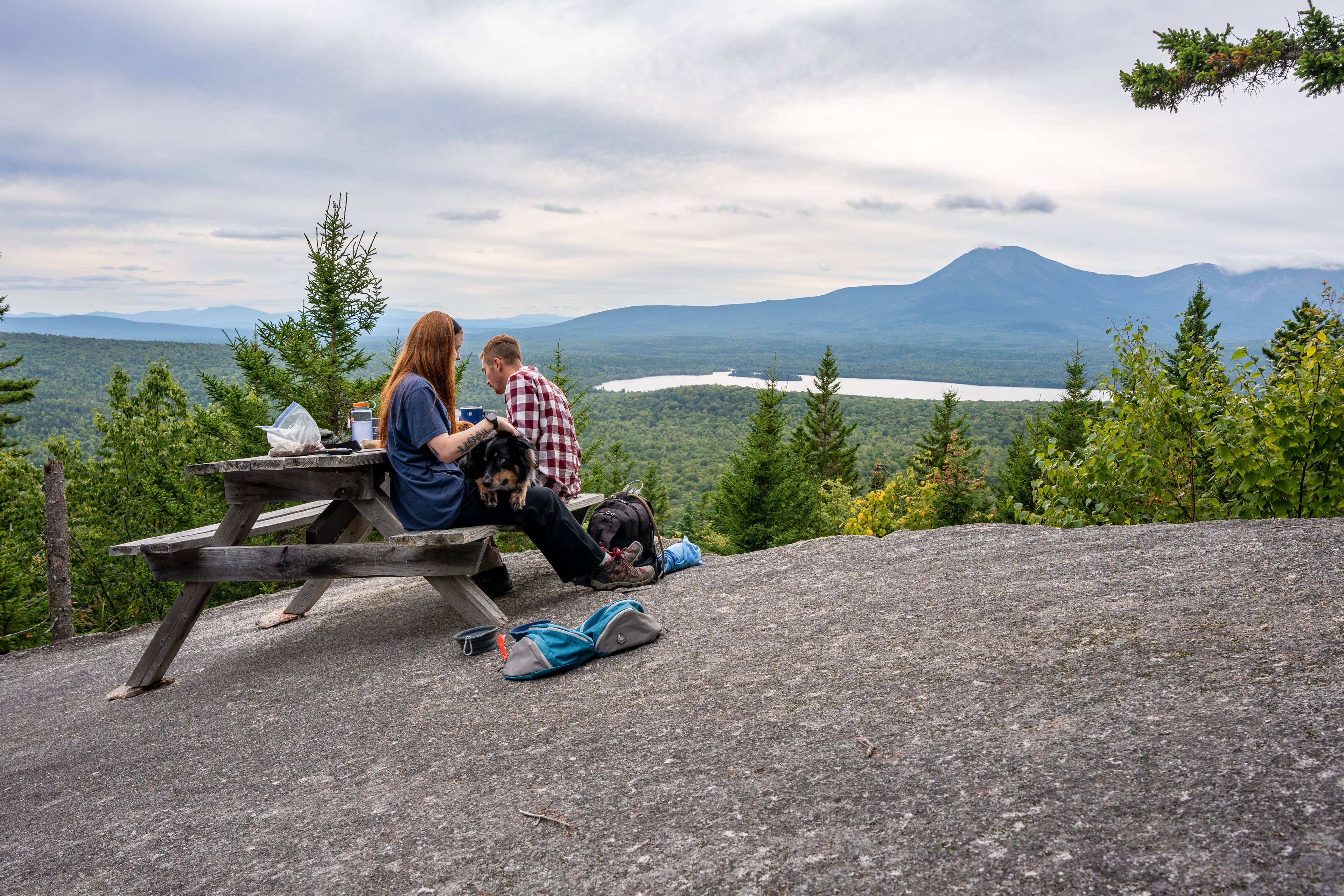 Two hikers and their dog on a leash have a picnic with a view on top of Barnard Mountain.
