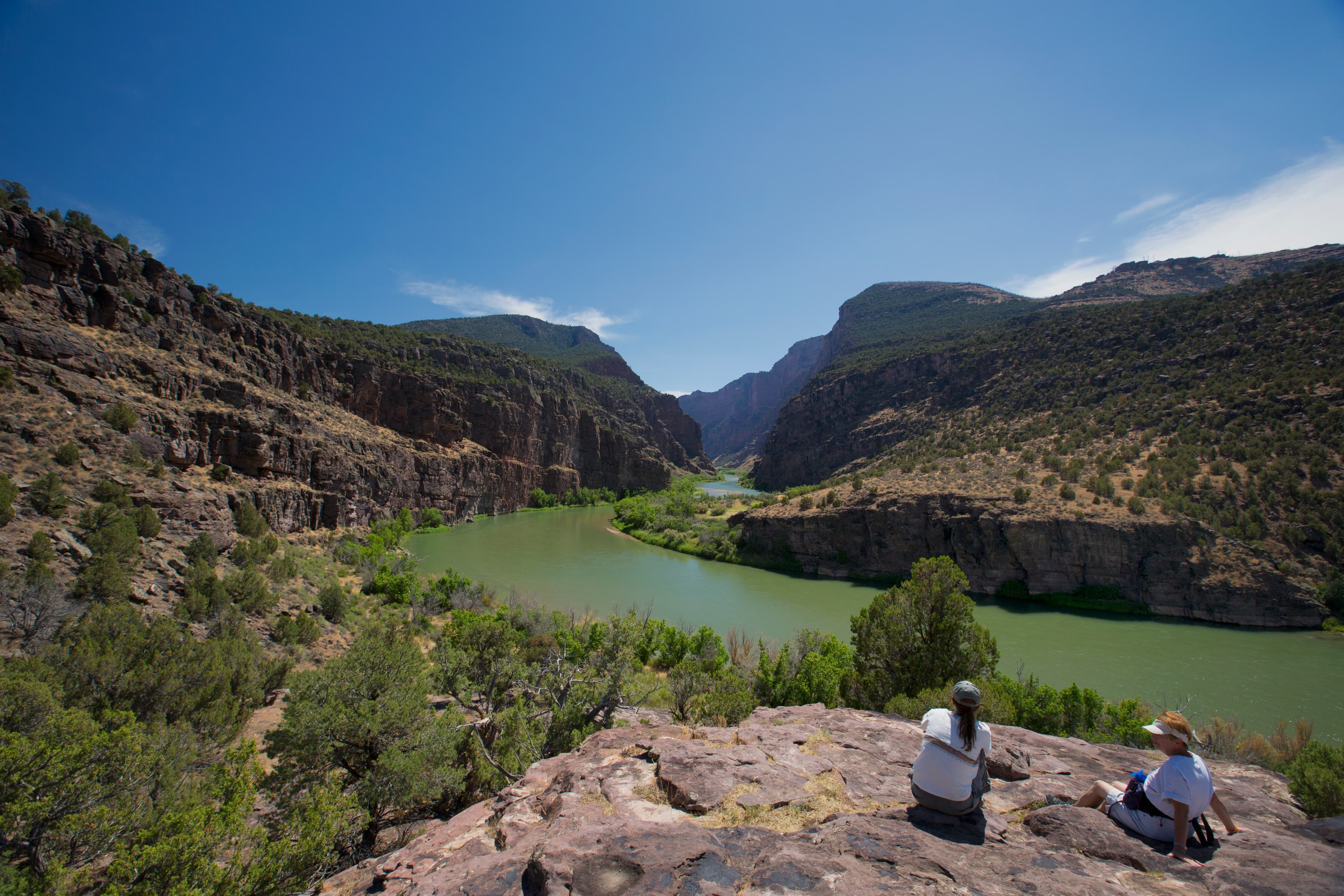 Two women sit on a large rock as a green colored river flows into a canyon in the distance.