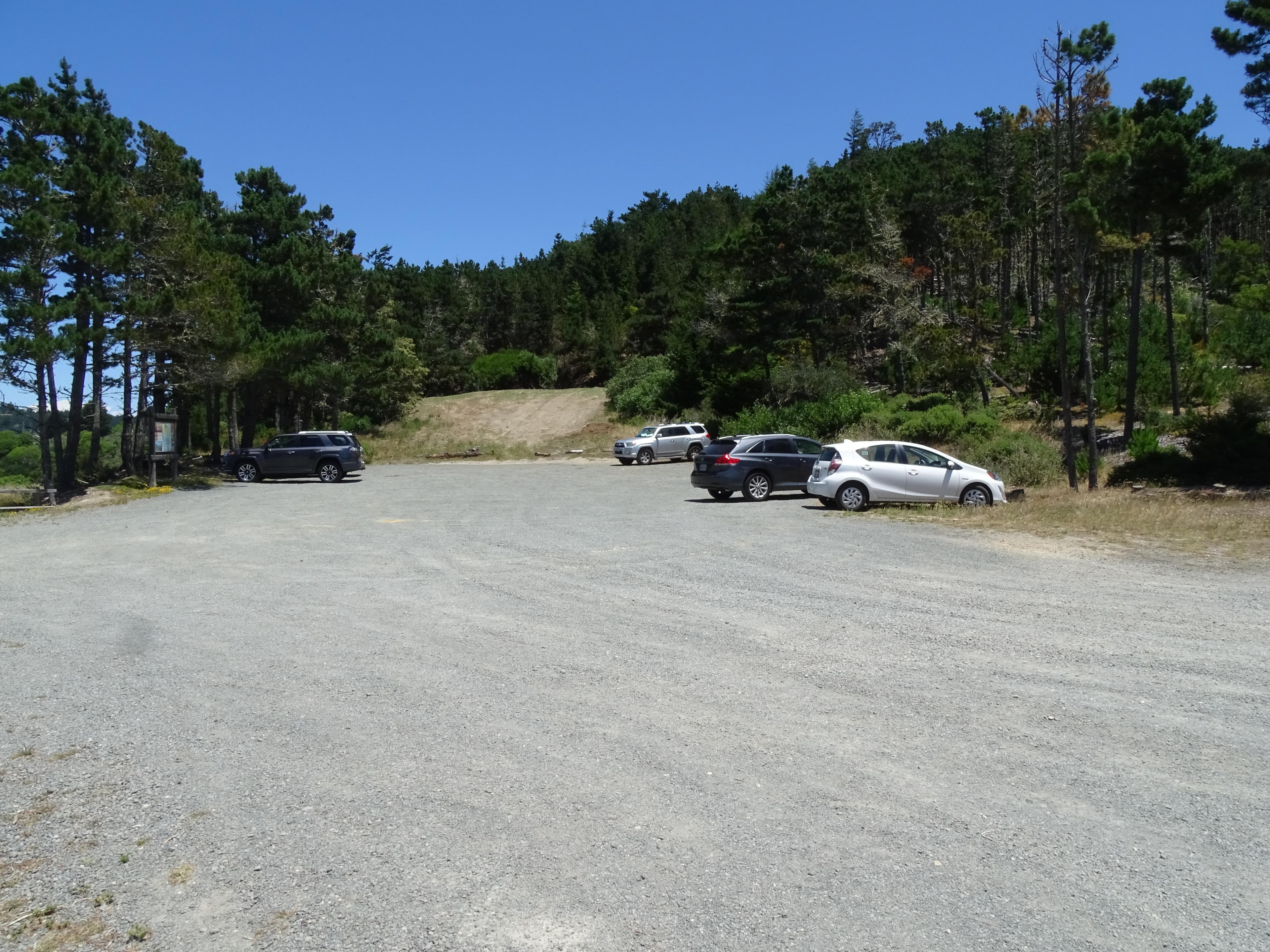 Five vehicles in a small gravel parking lot bordered by trees and a large brown sign board.