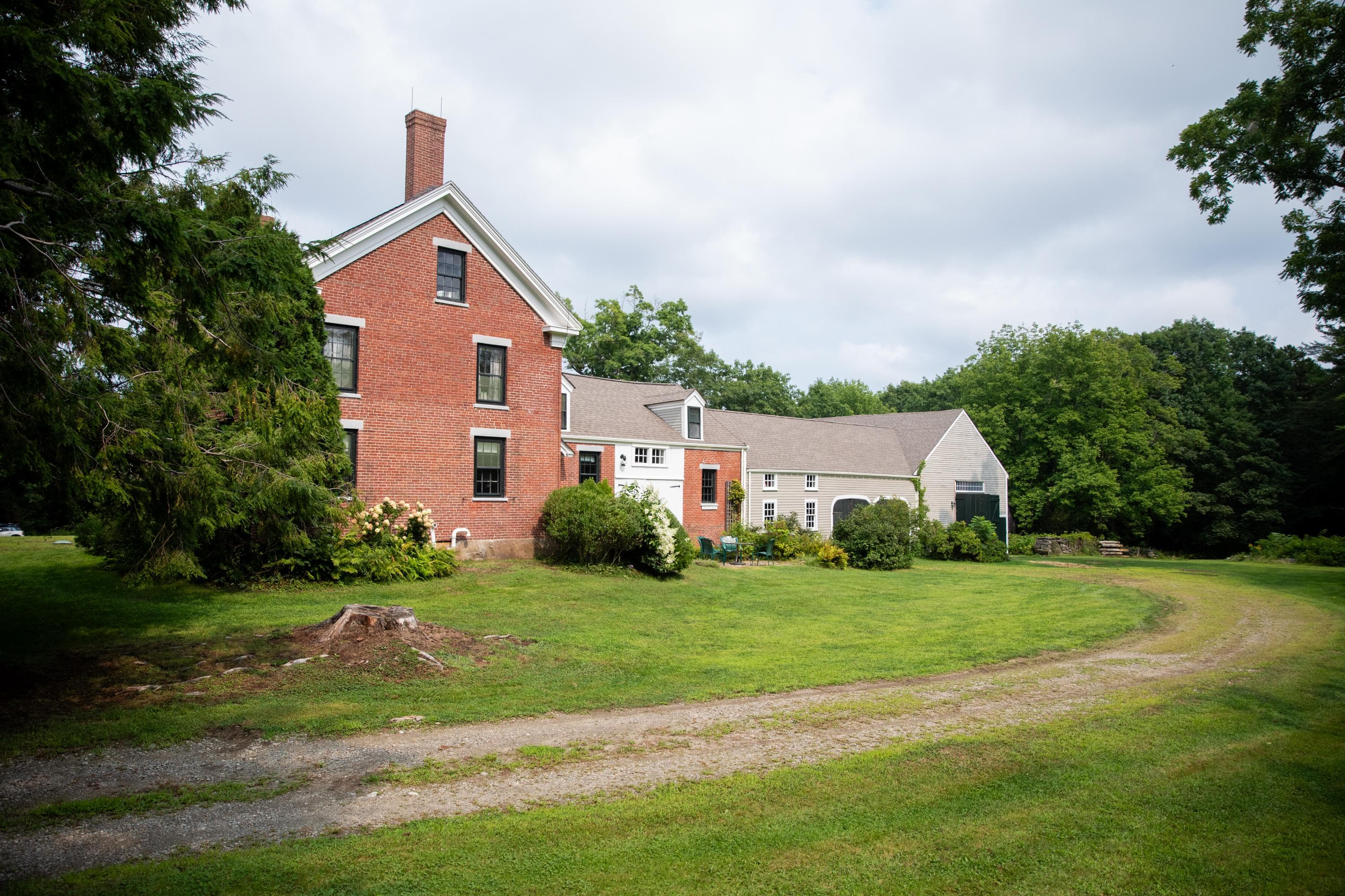 A sandy brown driveway curves through green grass towards the two story-red brick homestead and grey