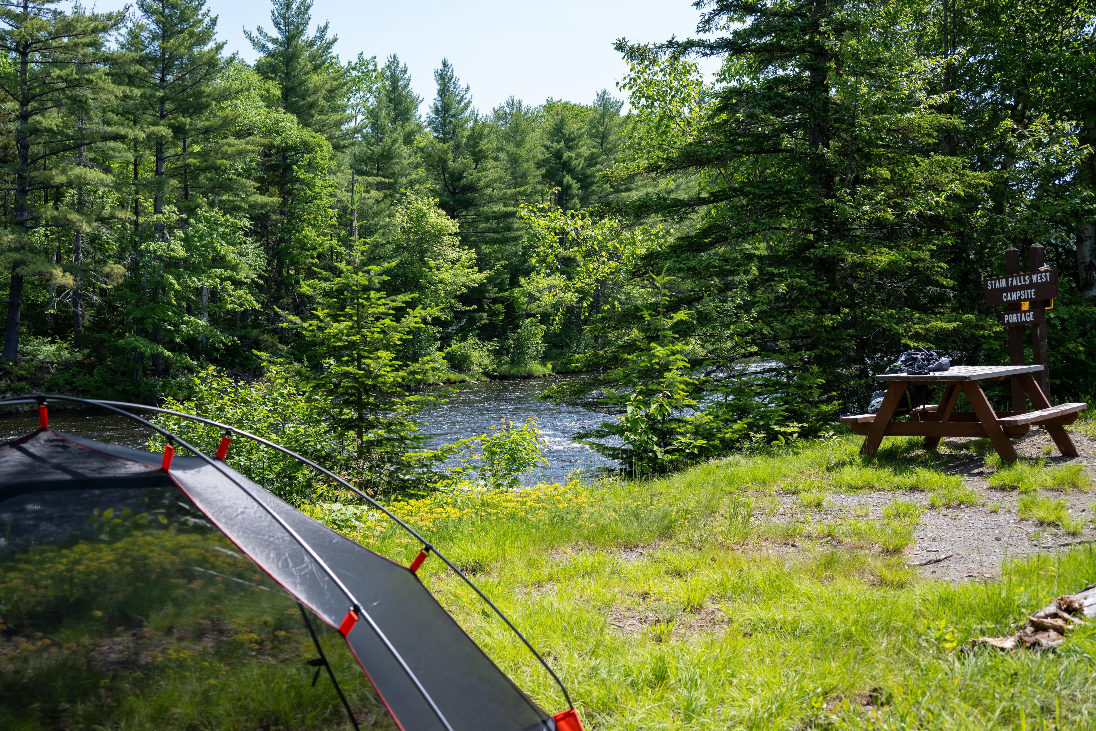A tent at a grassy campsite area with a picnic table and fire pit next to a river on a sunny day.