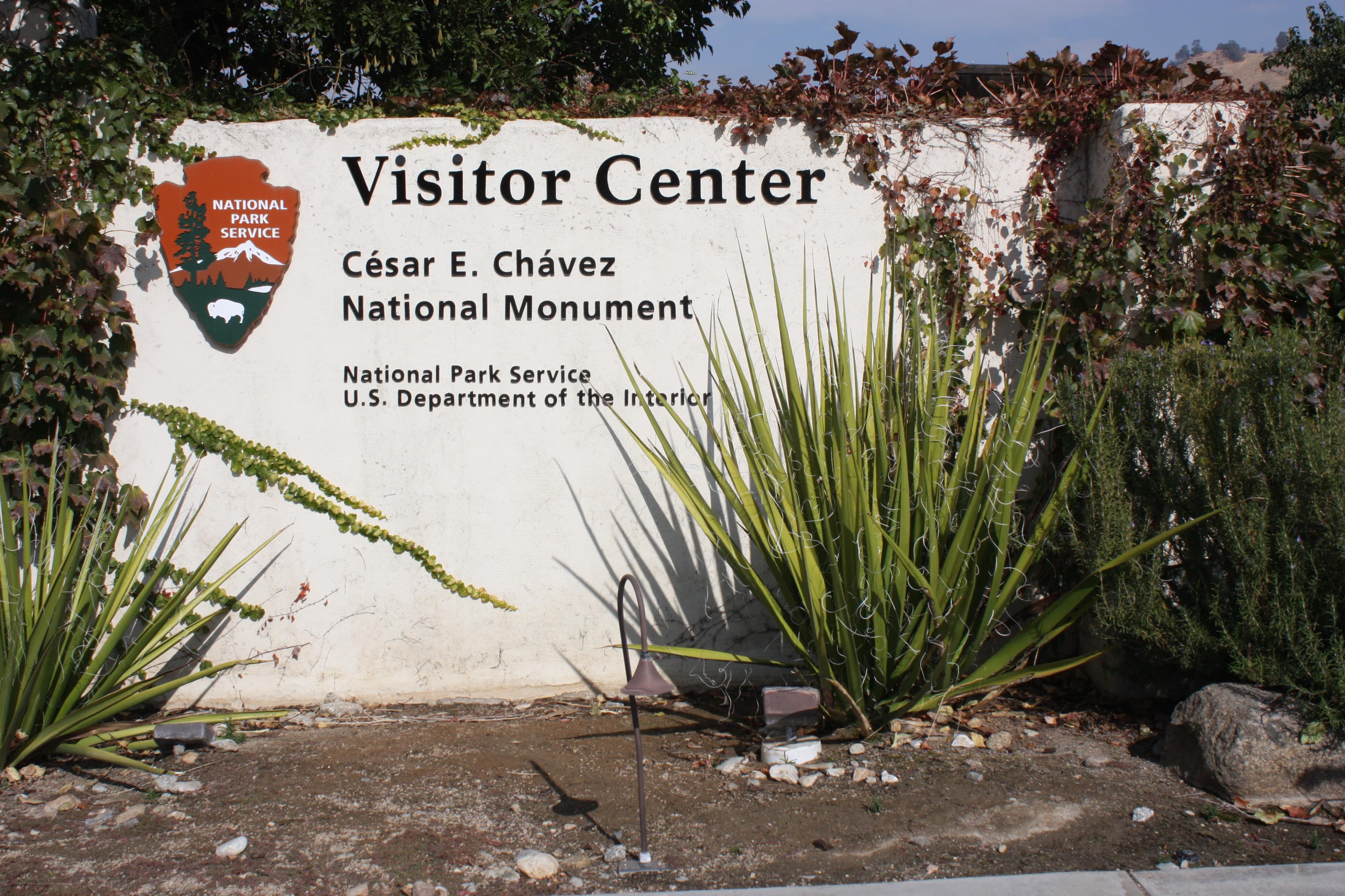 A white stucco wall featuring the Park Service arrowhead symbol and words. Plants in the foreground.