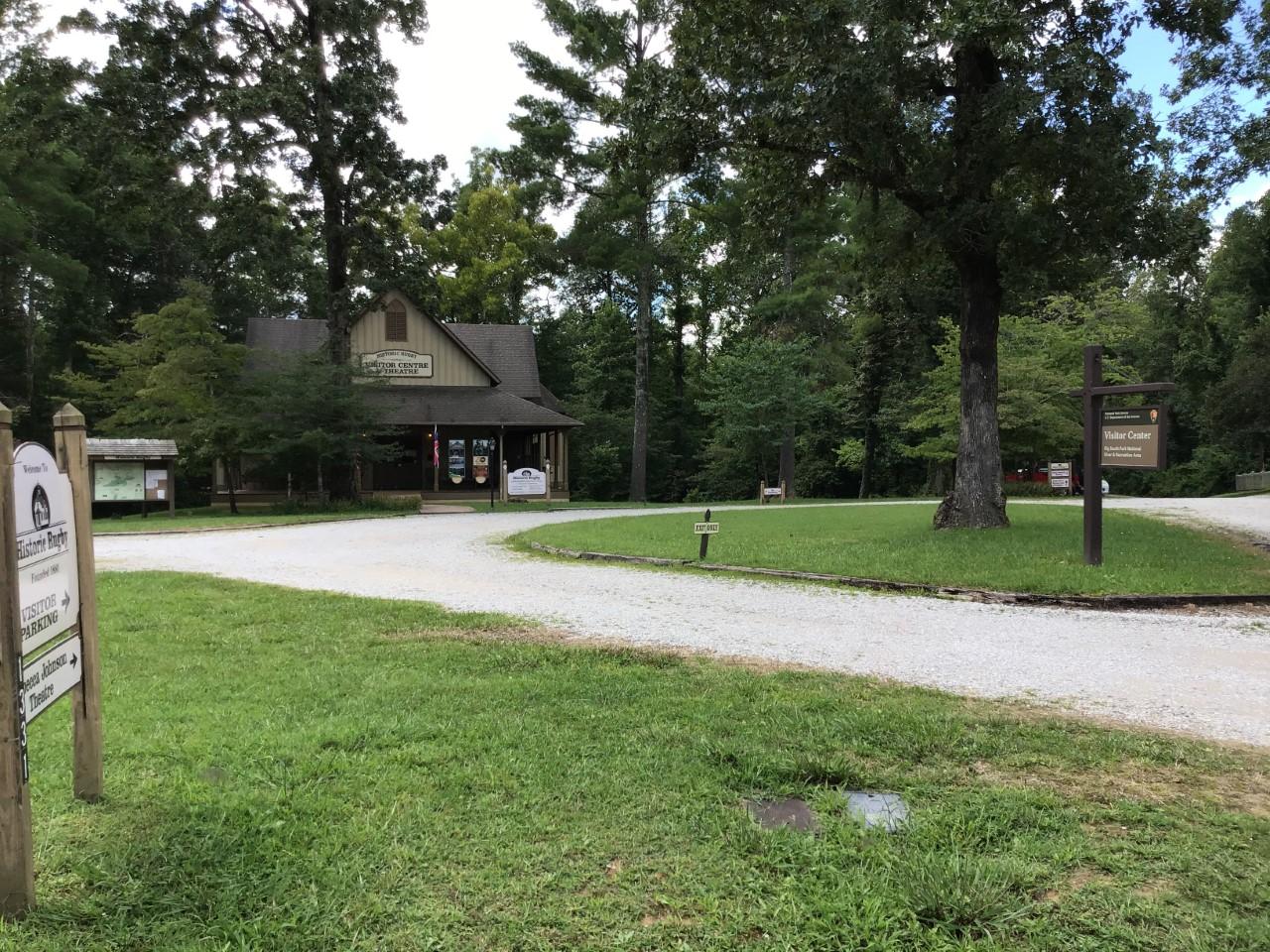 Tall trees surround a small building with a gravel parking lot in front.