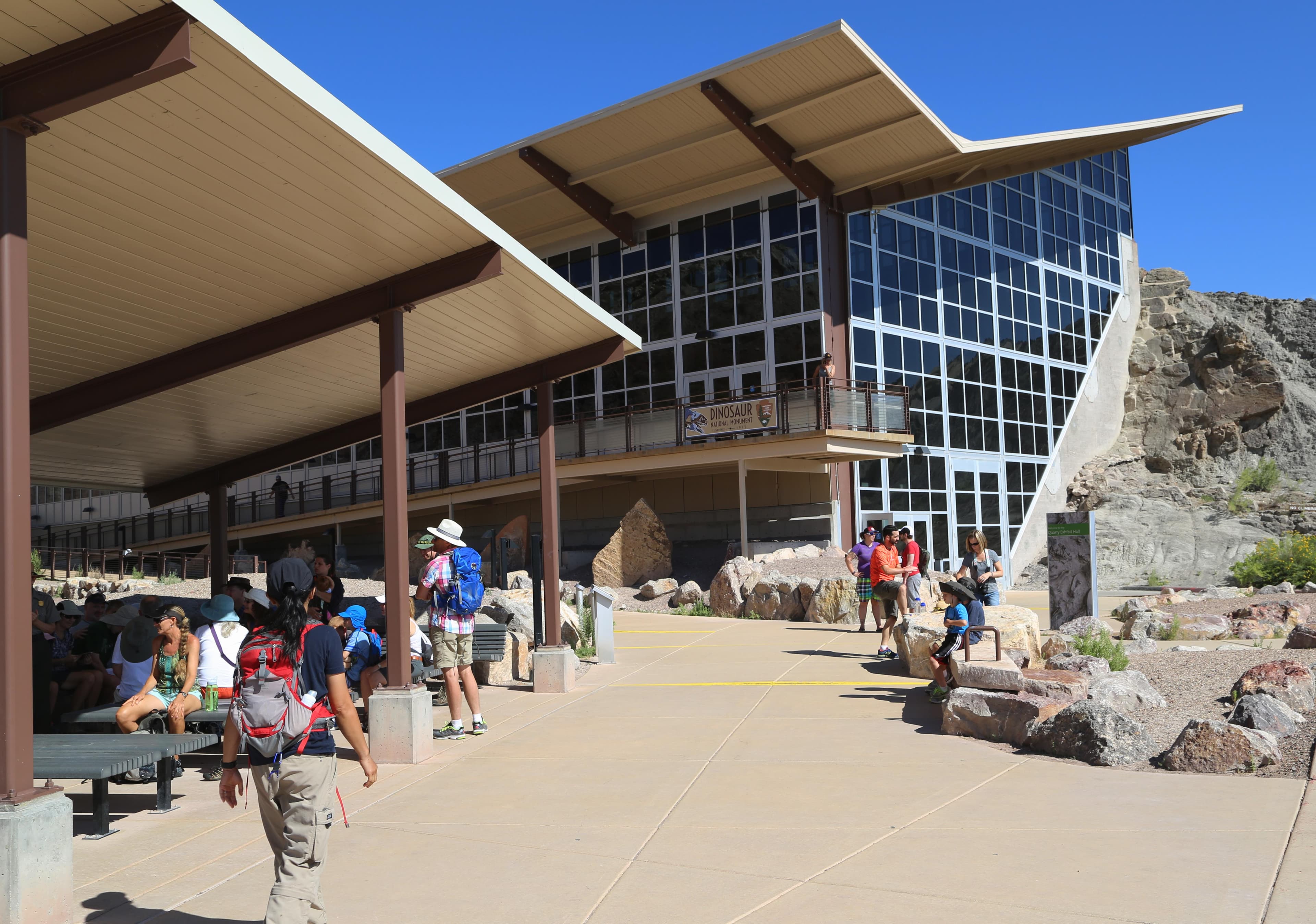 People stand and walk on a sidewalk in front of a large glass building with and angular roof