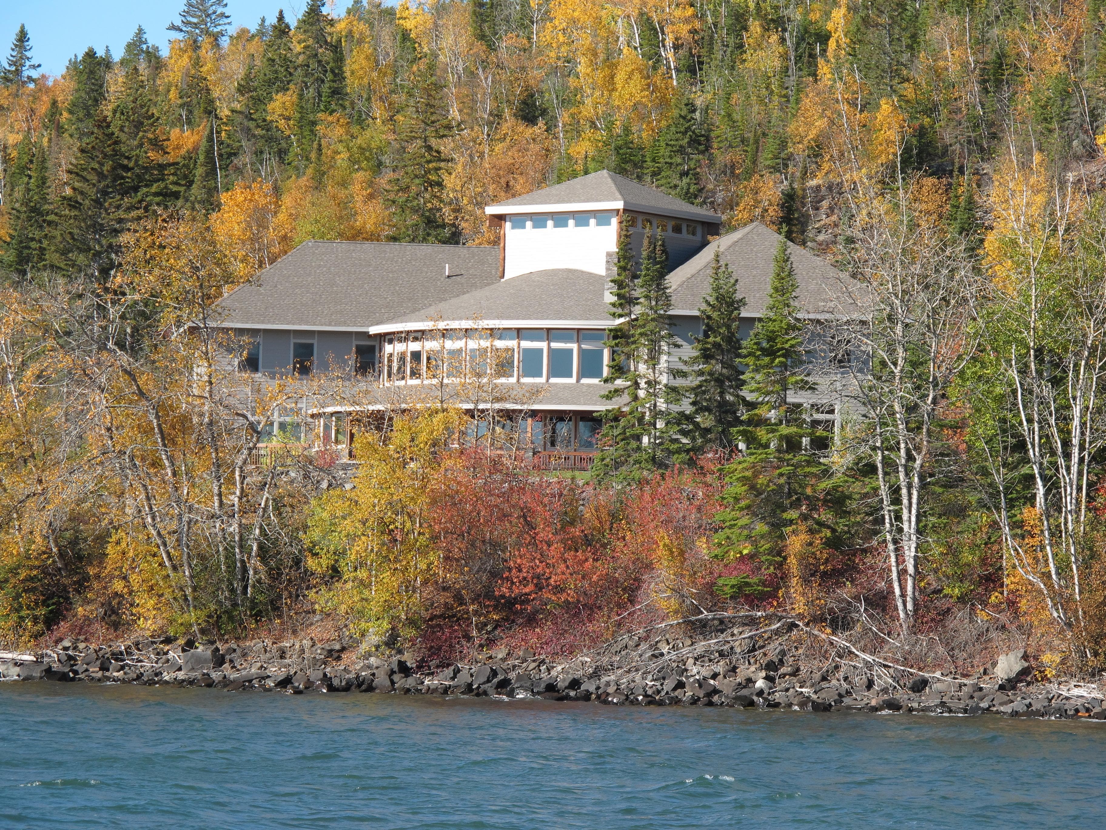 A large building with rows of windows nestled in fall colored trees and water in the foreground.