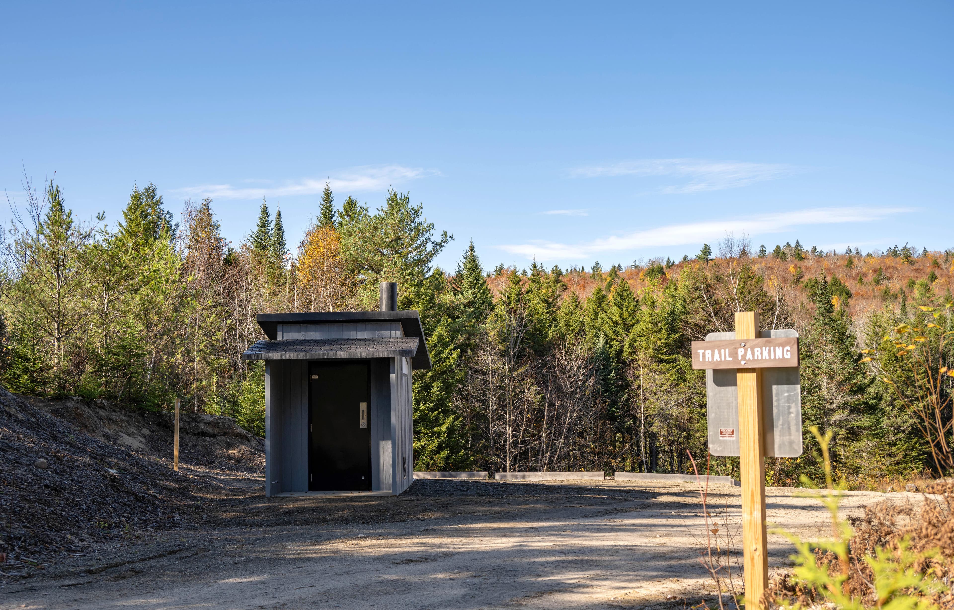 A dirt and gravel parking area with a vault toilet to the left side on a sunny fall day.
