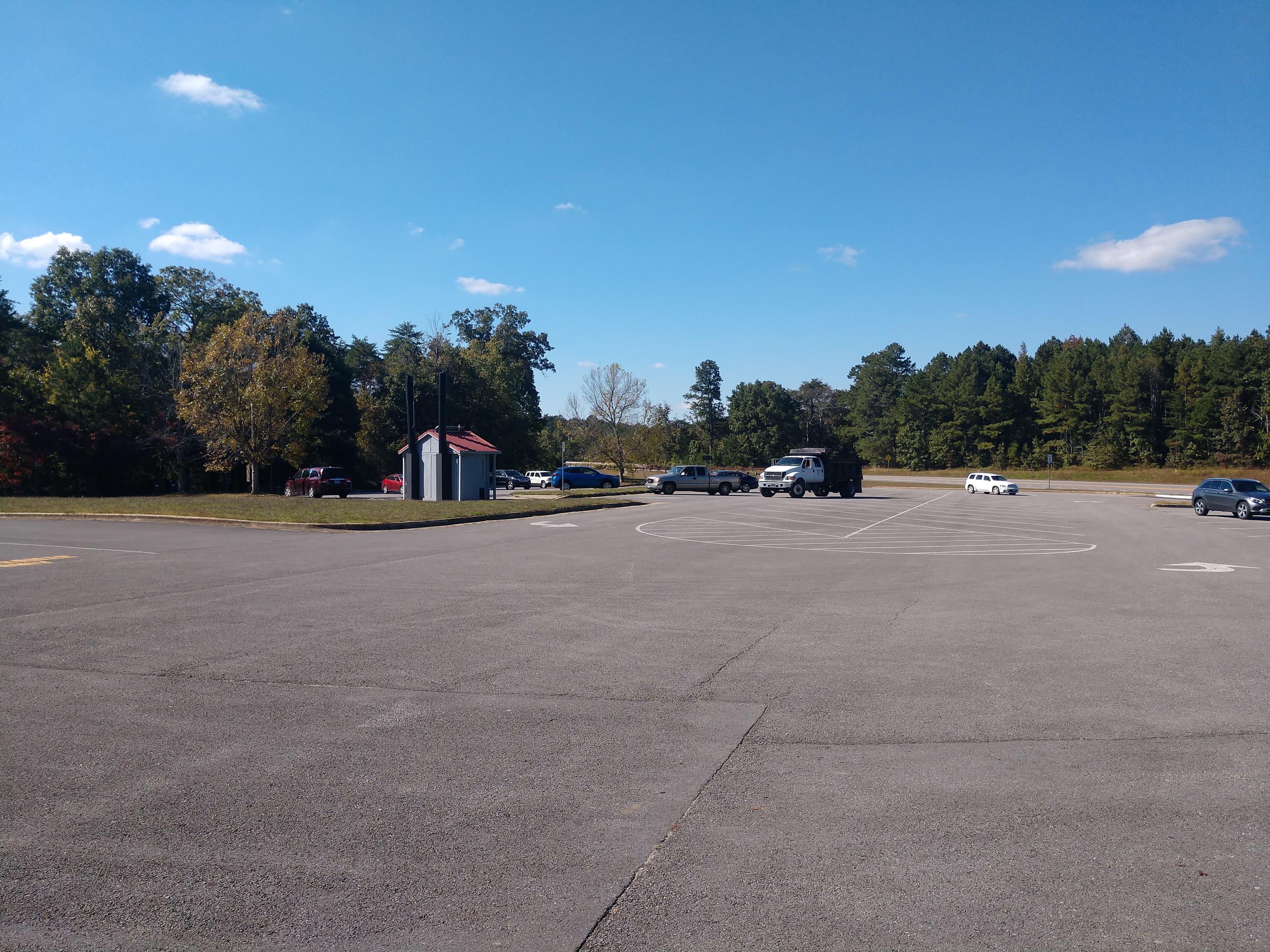 A large paved parking area surrounded by trees with a small vault toilet facility in the middle.