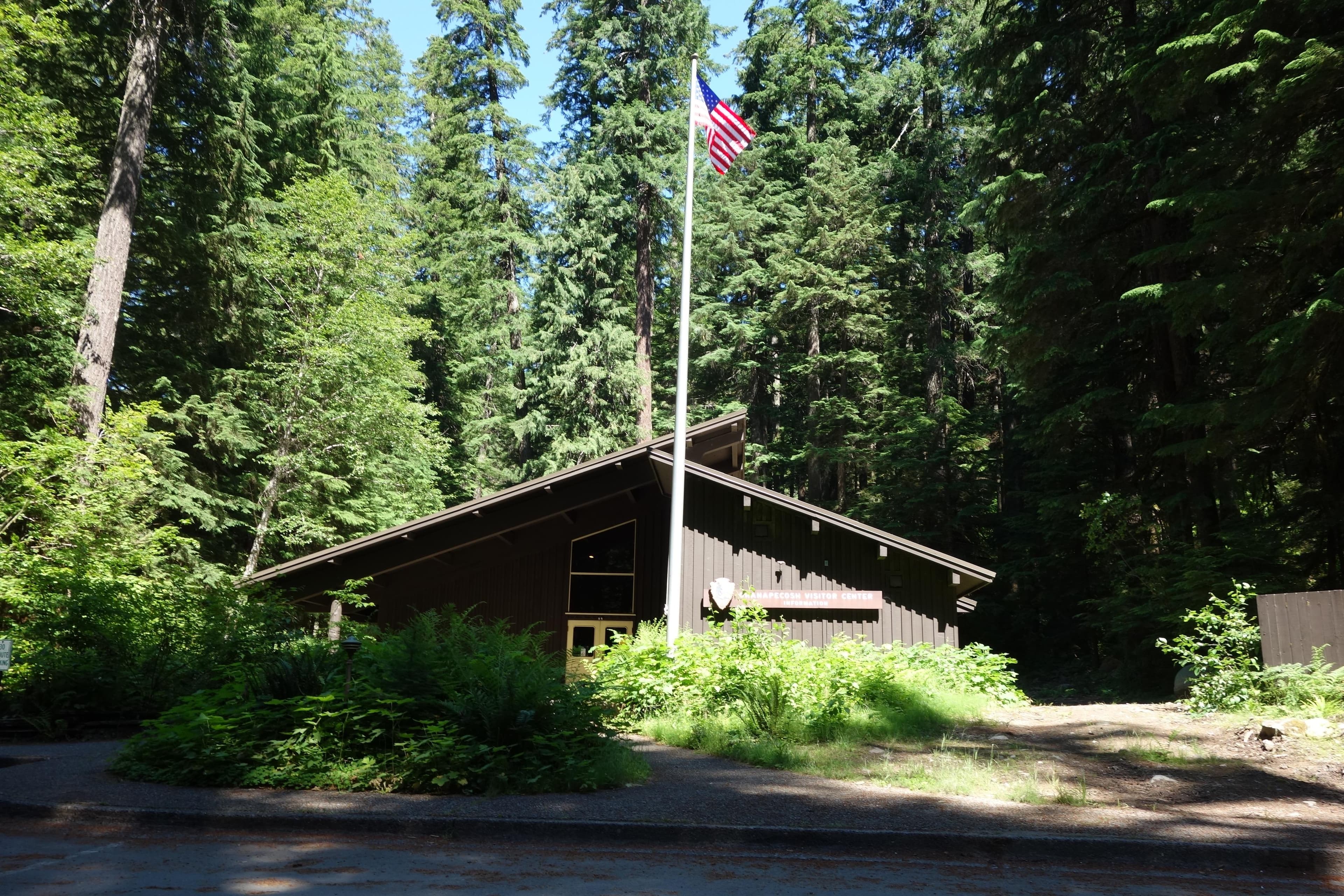 A brown building with a pitched roof sits surrounded by tall old growth trees.