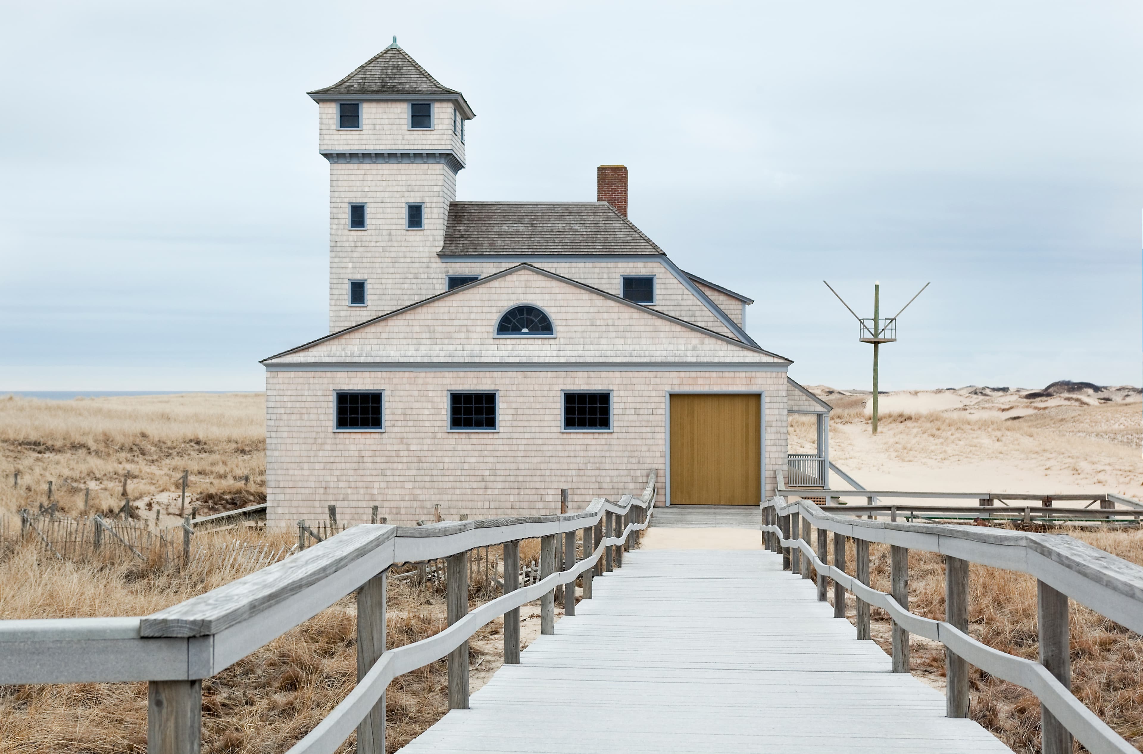 Ramp leads down to garage of Old Harbor Life-Saving Station. House against dunes and gray sky.