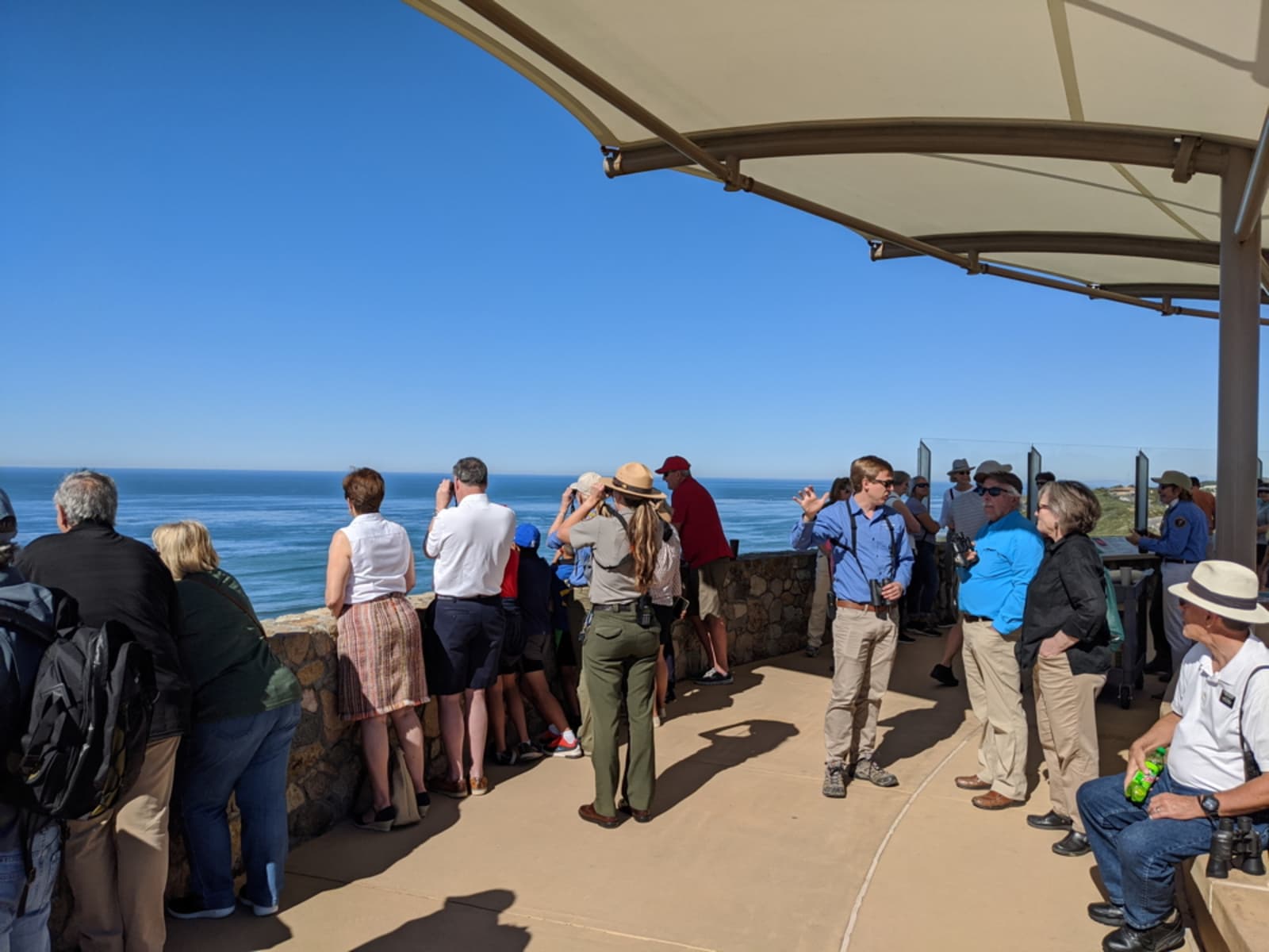 A crowd of people gather under a white, cloth covering. They look over a fence towards blue water.