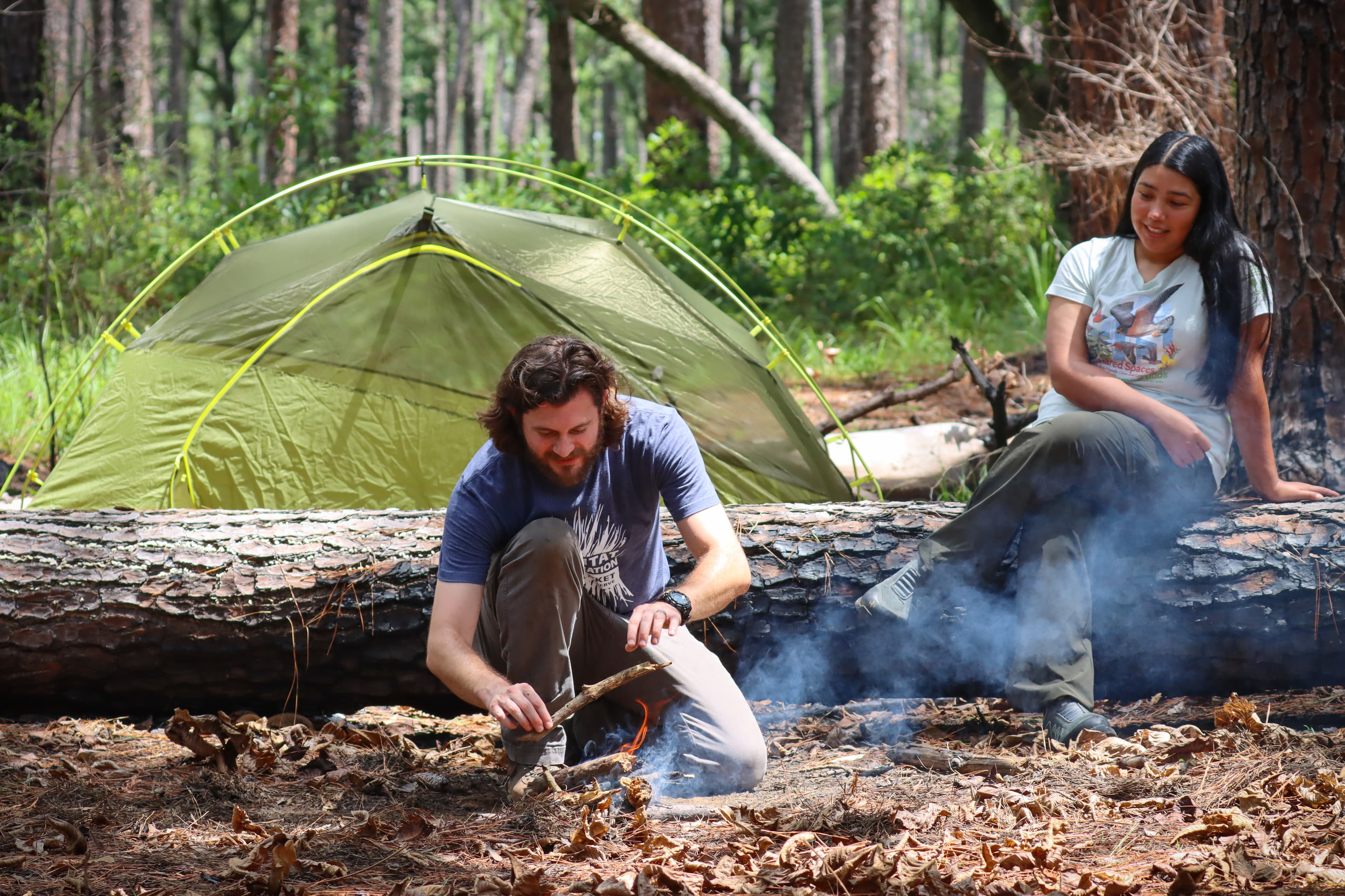 A camper builds a small fire while another camper sits on a fallen log with a tent in background.