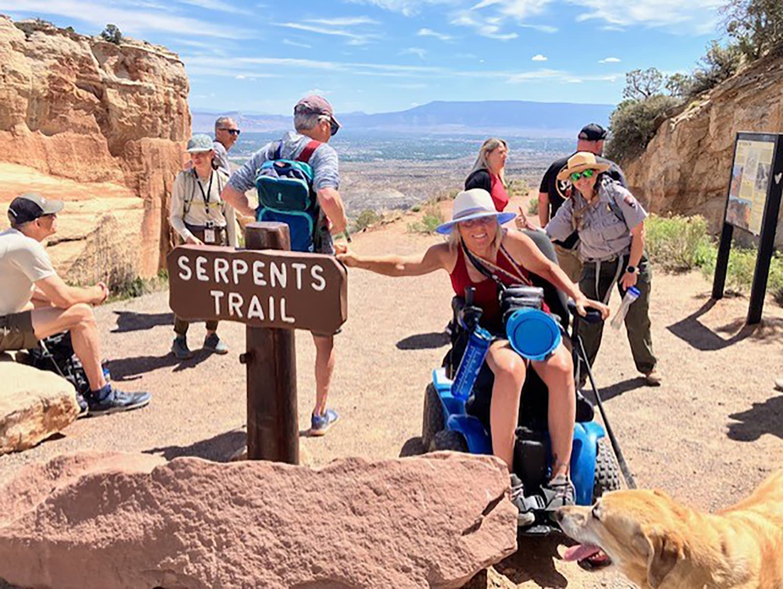 woman using wheelchair and service animal points to Serpents Trail sign with uniformed ranger behind