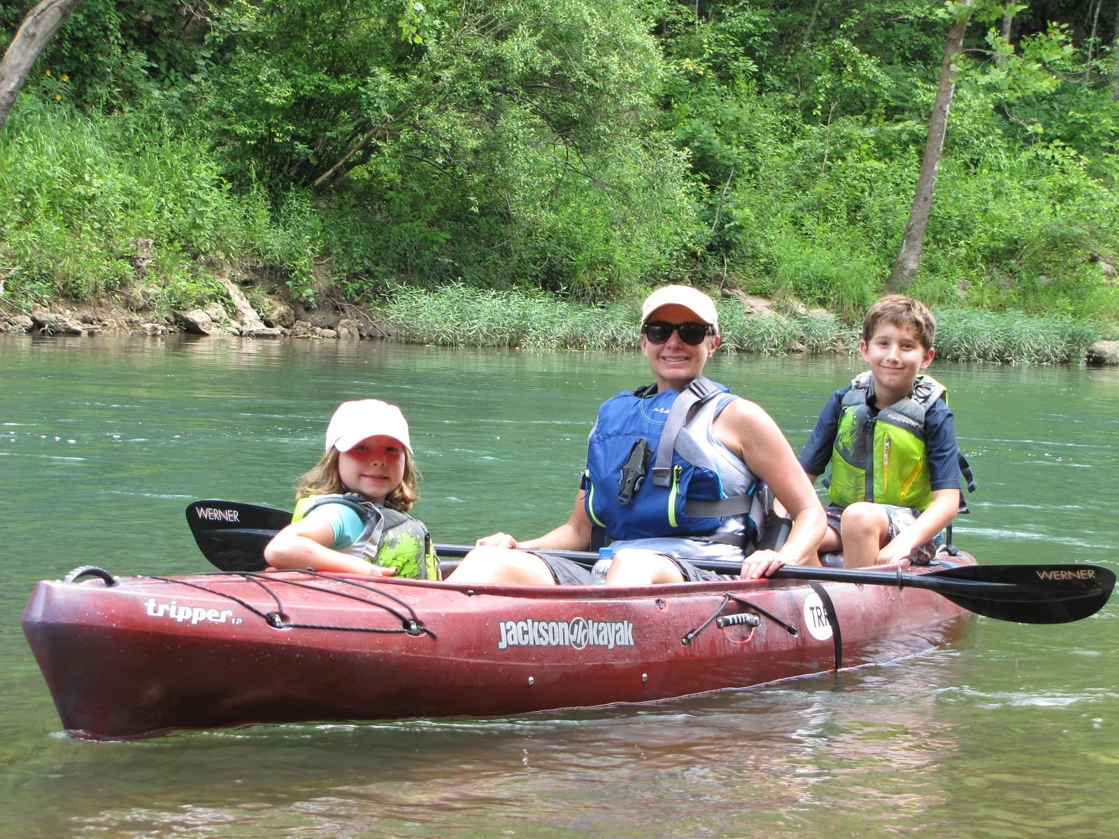 Red kayak on river with woman in middle, girl in front, boy in back, all wearing PFD
