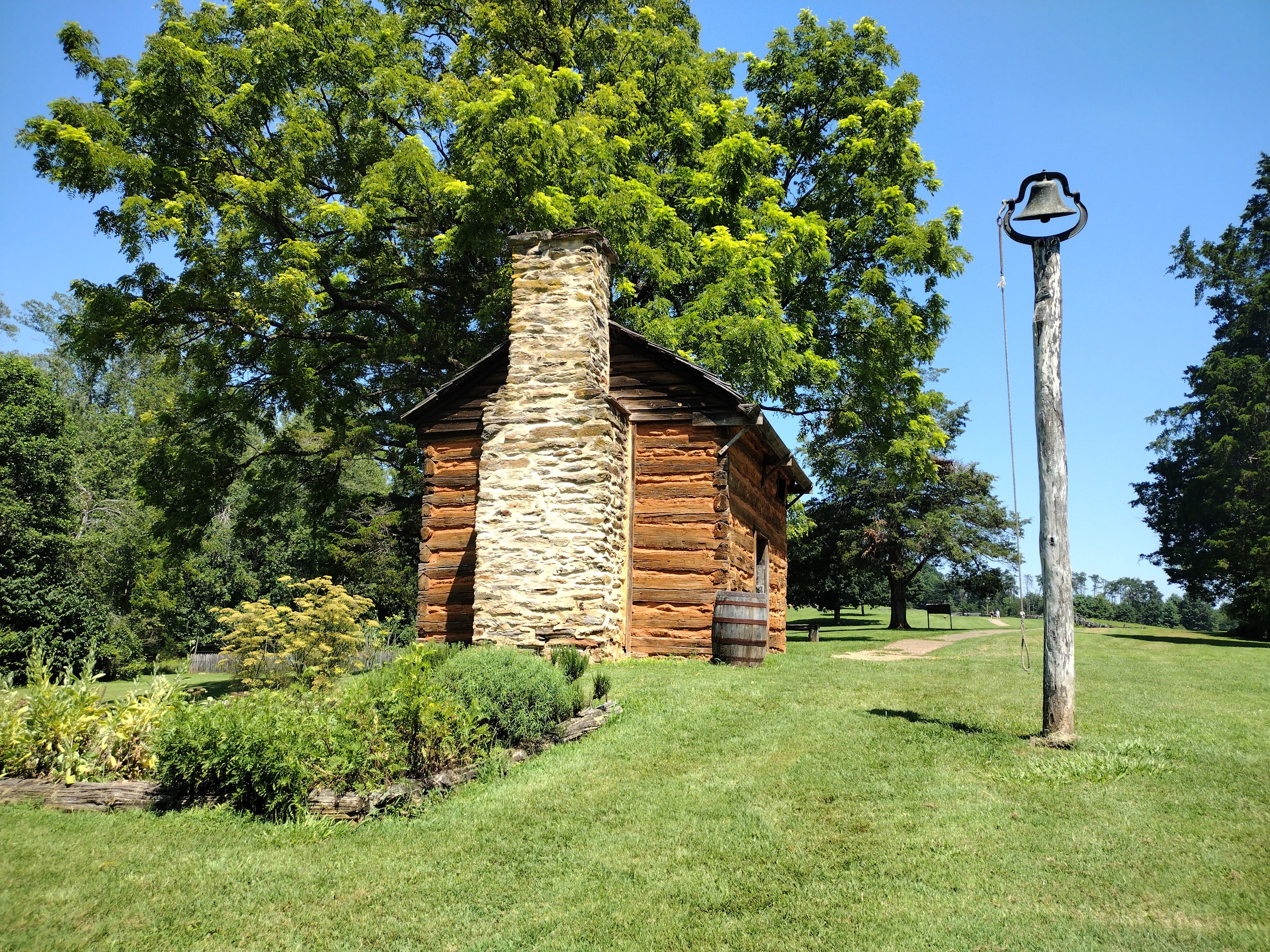 A rustic cabin with a stone chimney and a bell placed on a wooden post.