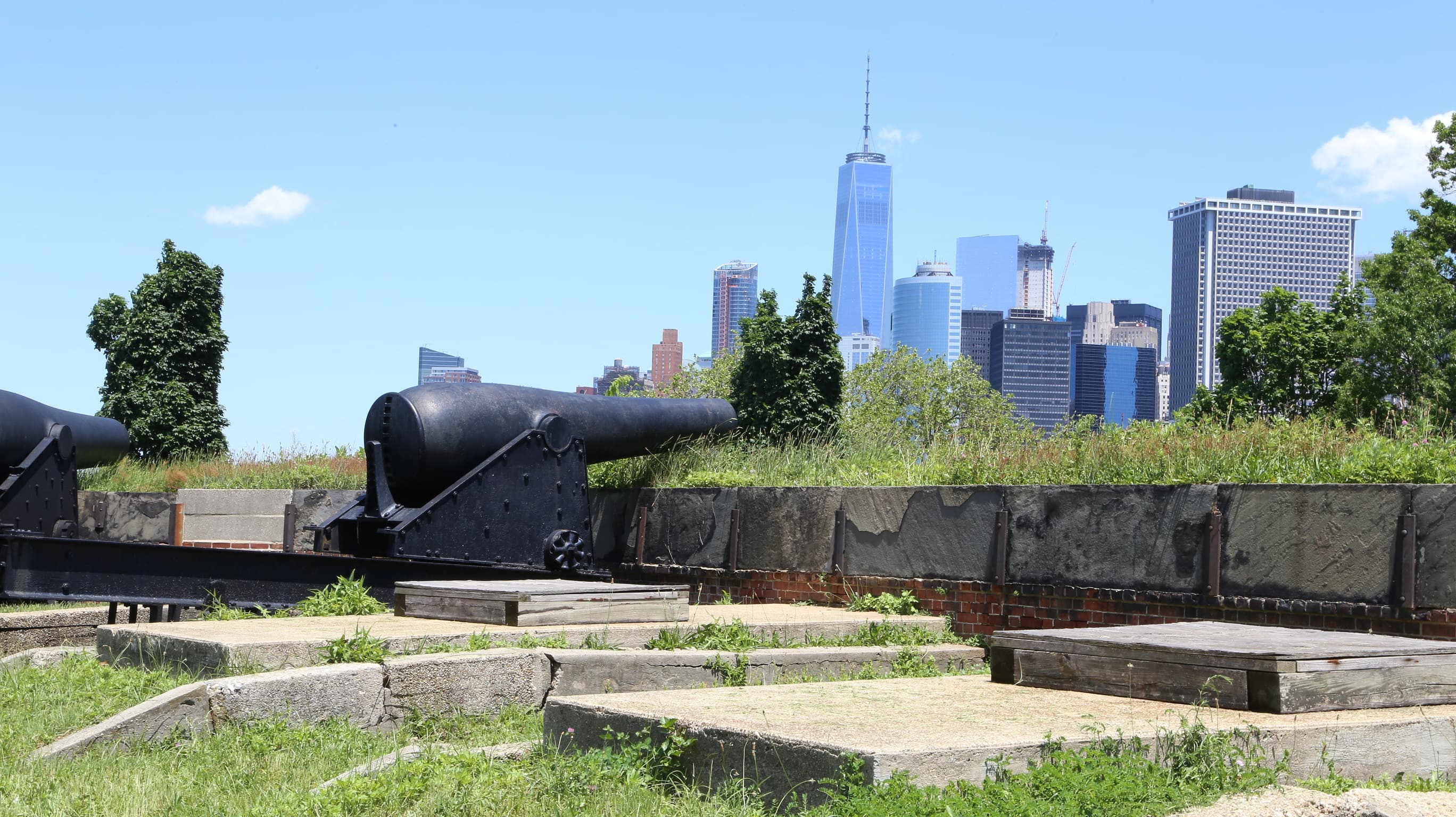 large, black, iron cannon pointing over forts wall with NYC skyline in backround
