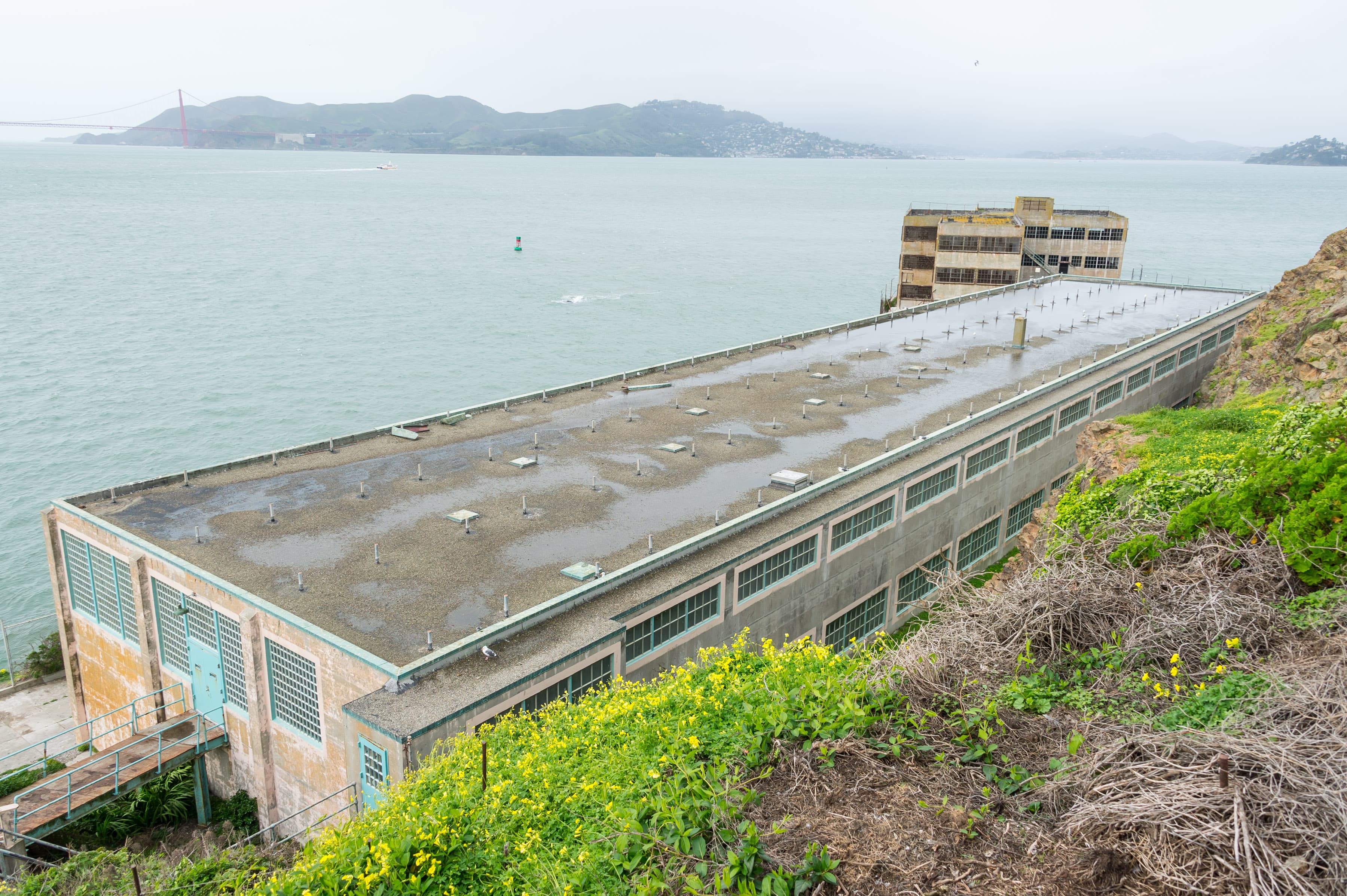 A long building next to a rocky plant covered slope, sitting in front of a large body of water.