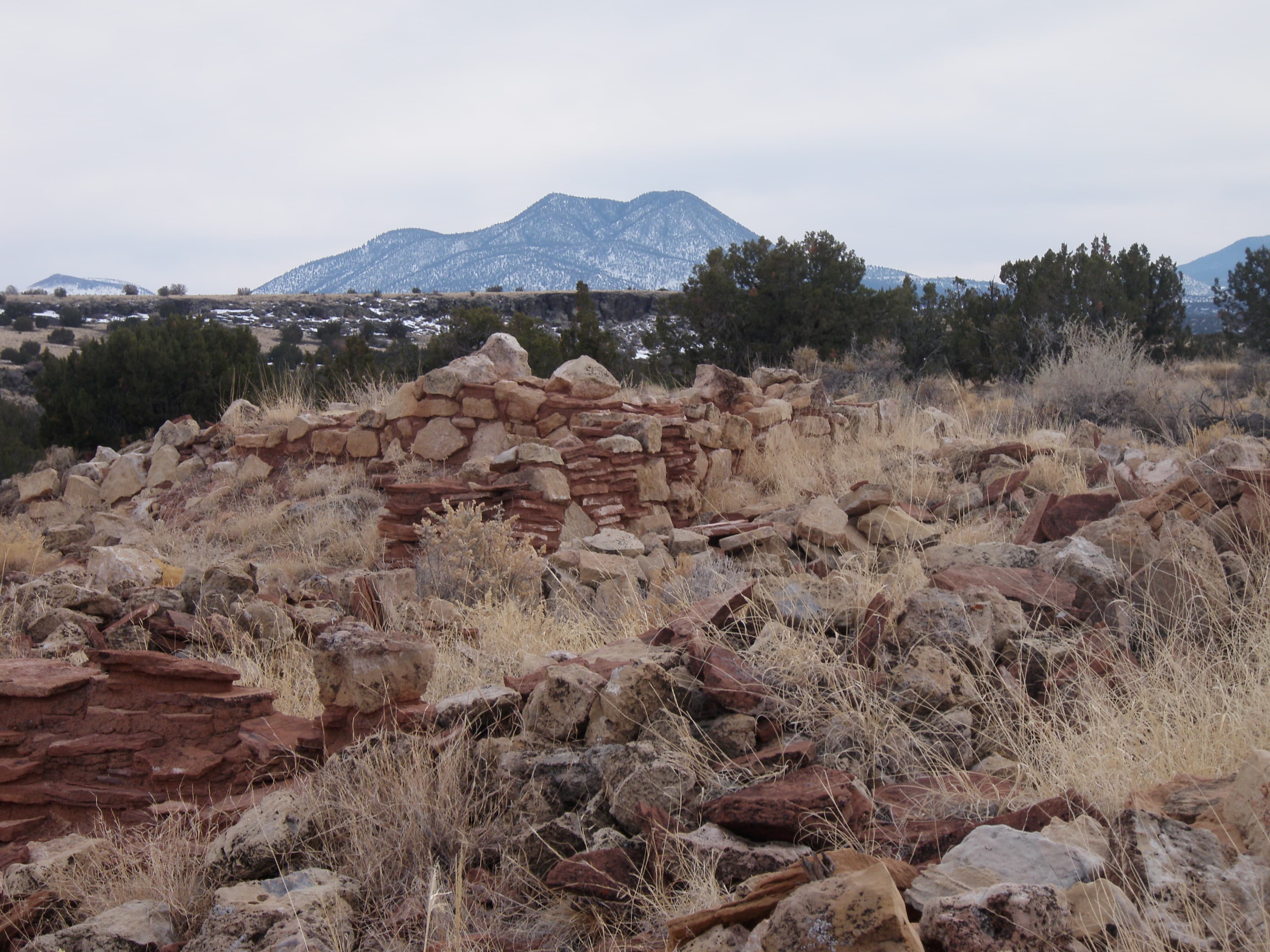 The remnants of a tan stone pueblo structure surrounded by yellow grasses and juniper trees.