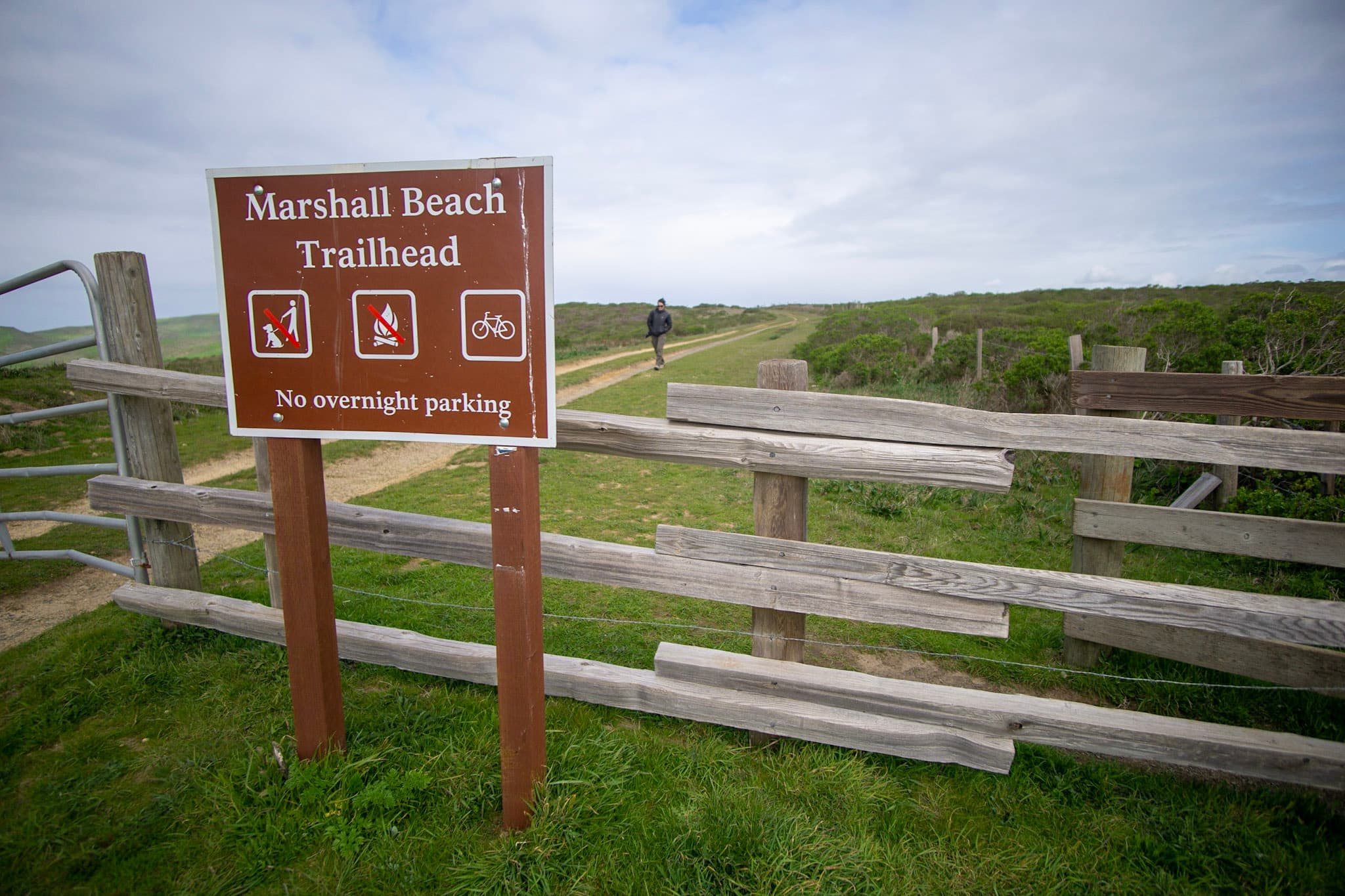 A brown trailhead sign alongside a split rail fence with a hiker on a broad trail in the background.