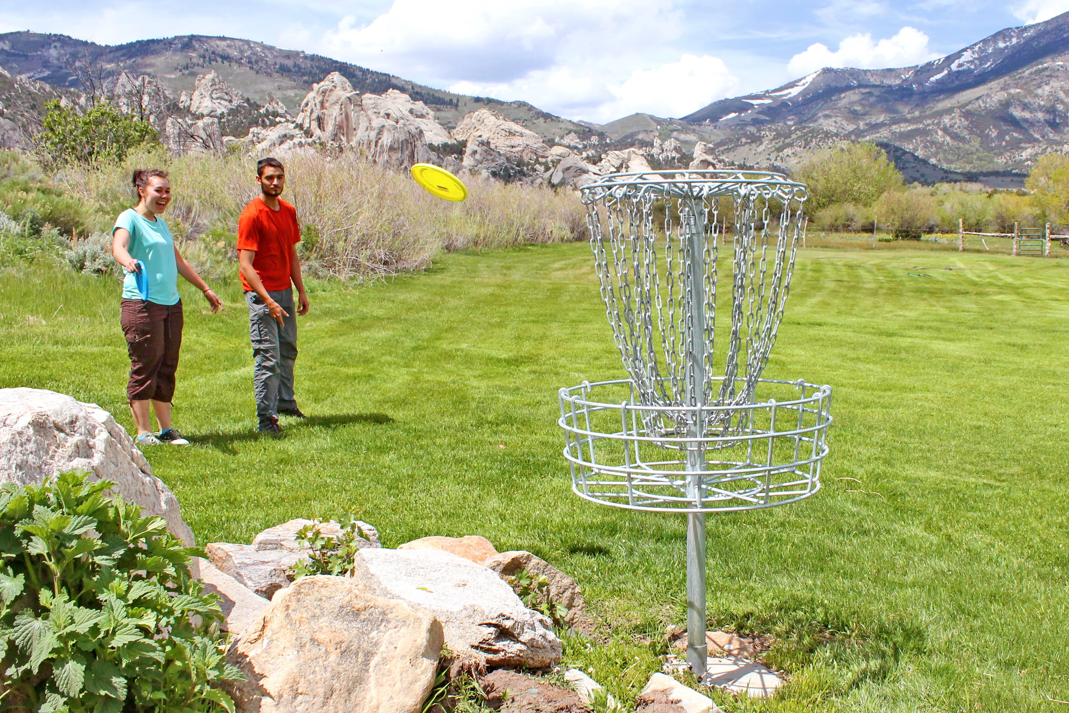 Two people watch a yellow disc flying toward the metal disc golf basket, castle rocks in the back.