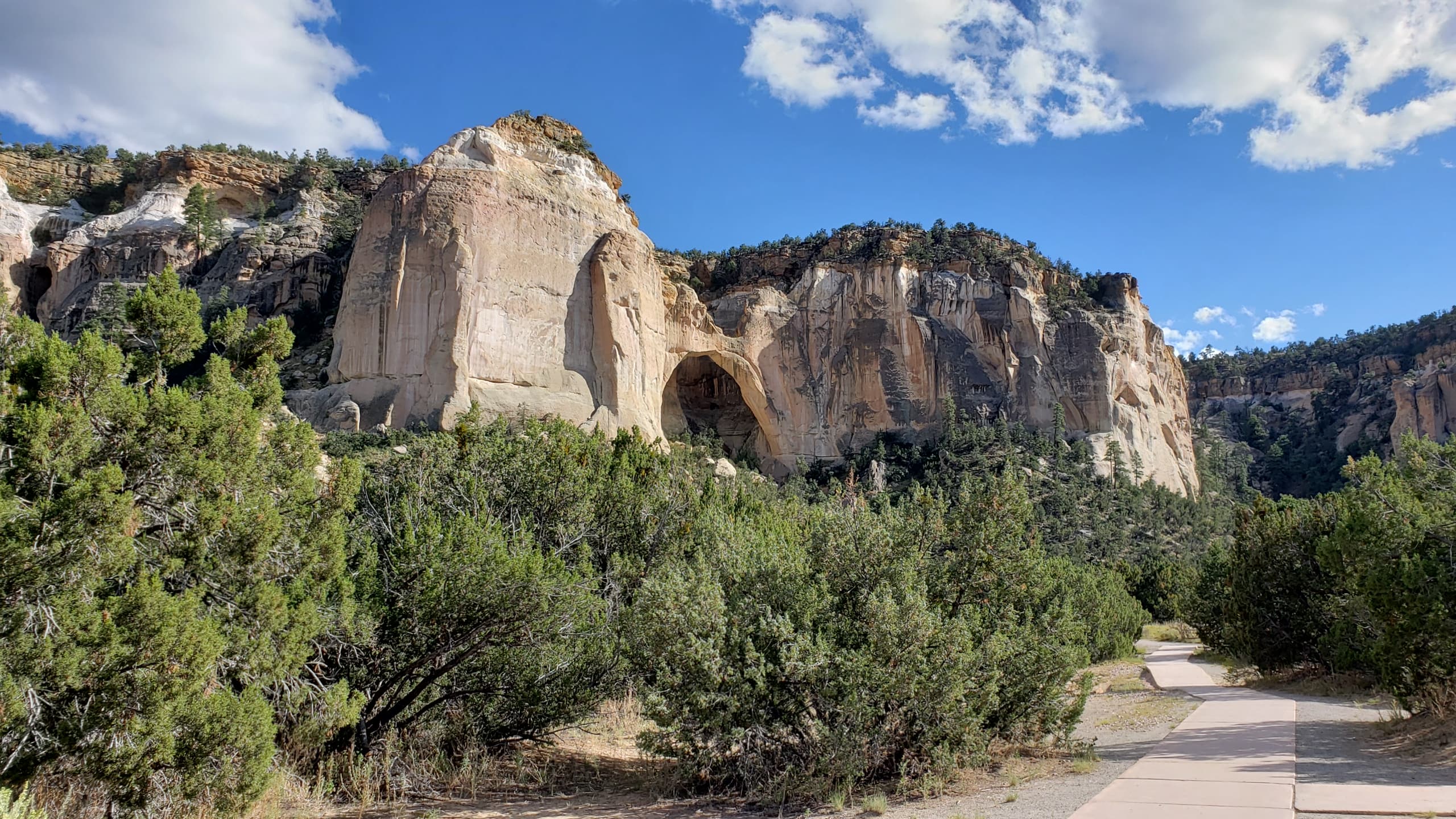 A sidwalk leads to a sandstone cliff with a prominent arch feature within the sandstone.