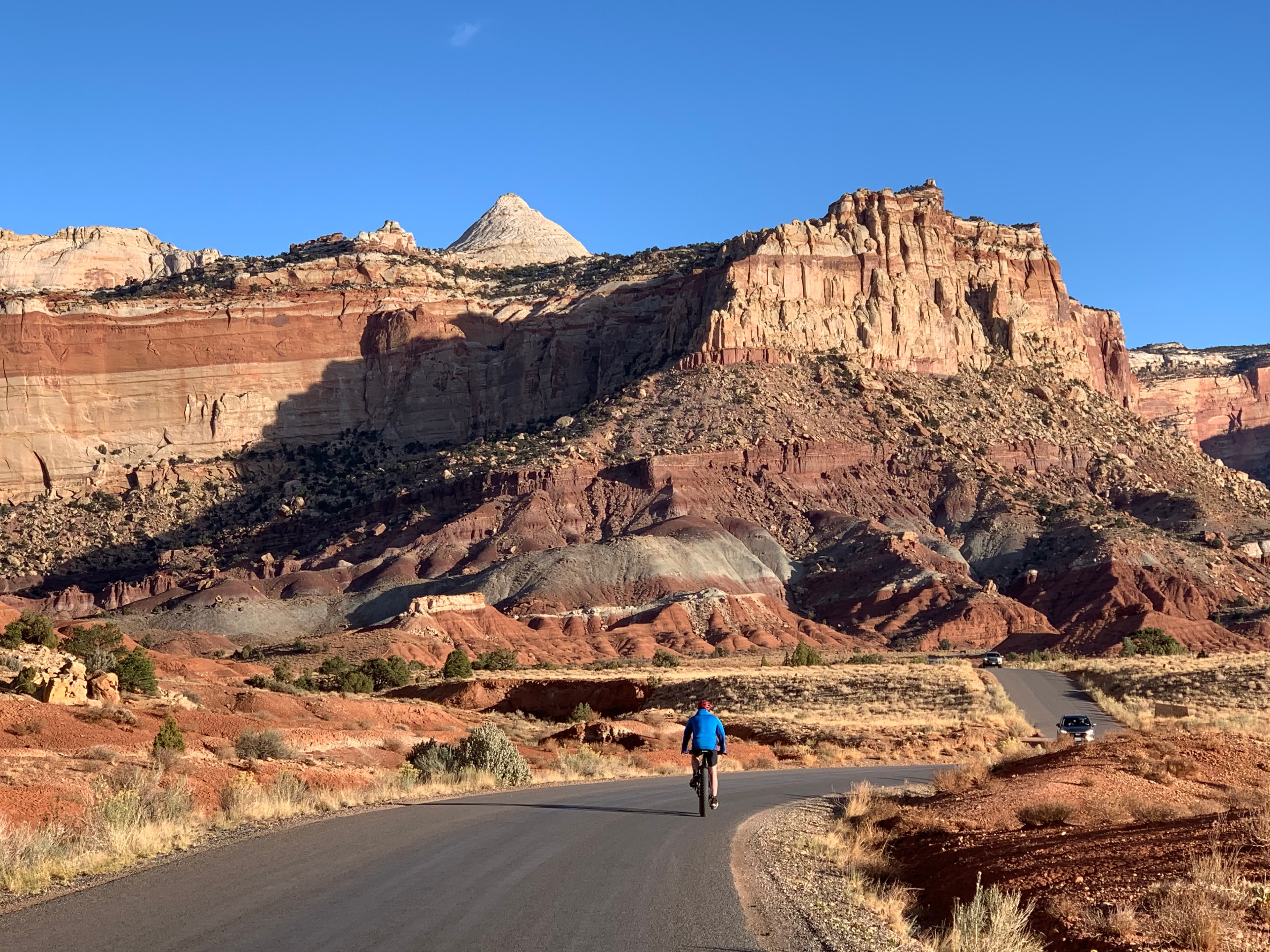 Person on bicycle on paved road with cars approaching and colorful cliffs above, and blue sky.