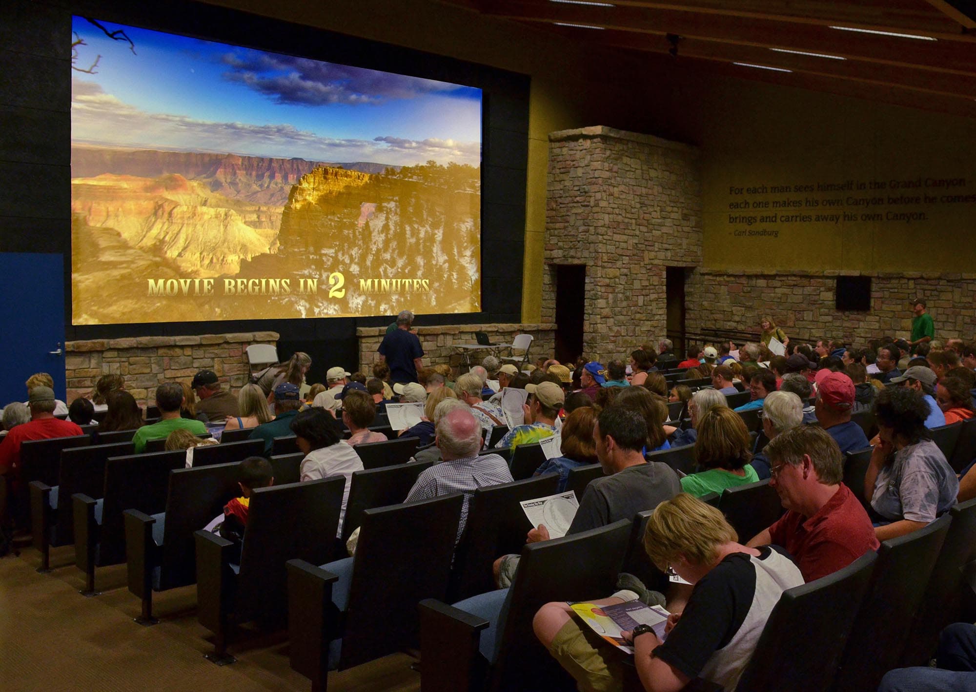 People are almost filling the seats in a darkened theater. Screen reads: Movie begins in 2 minutes.