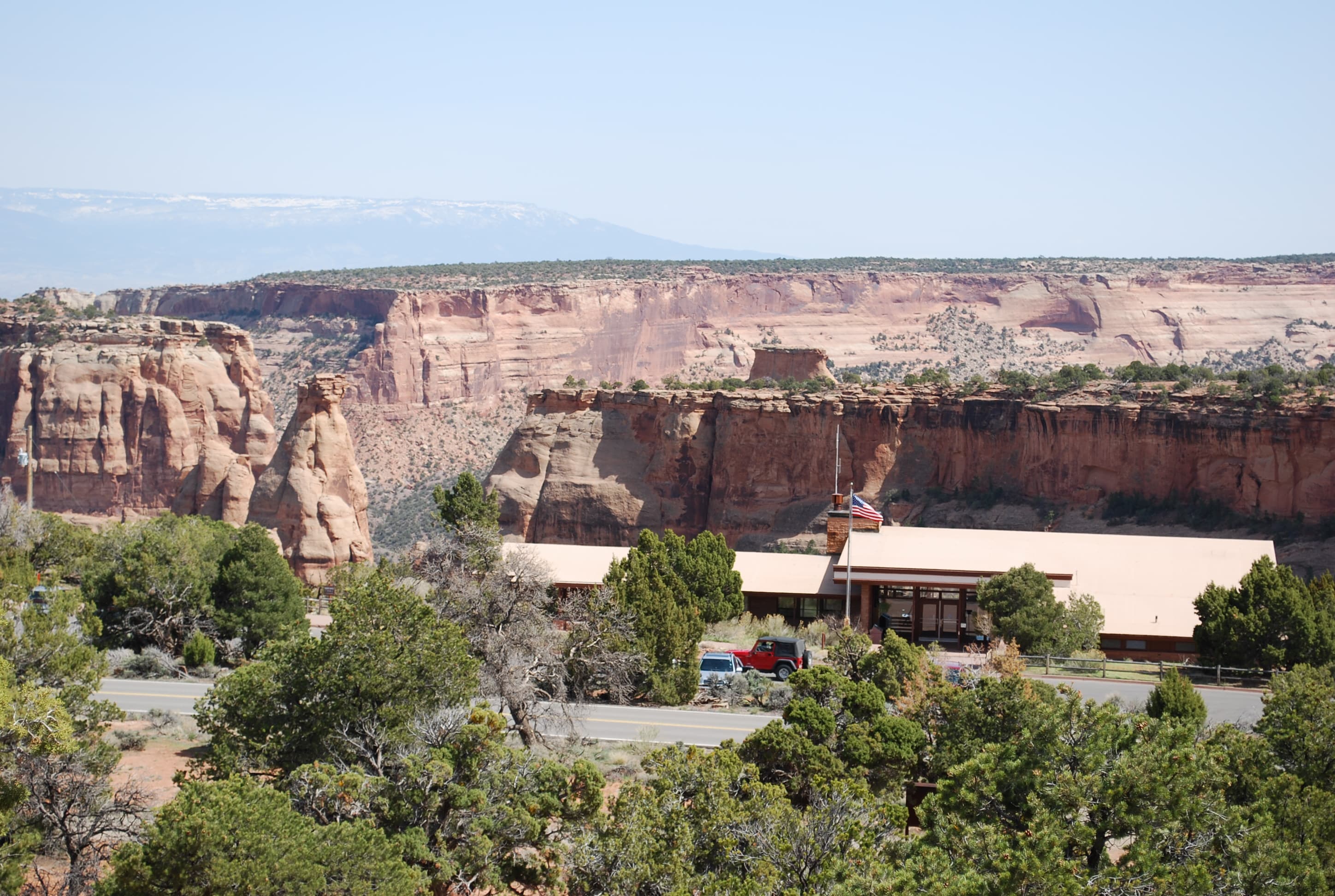A low, mostly flat building with a flagpole and shrubs, standing before a desert canyon