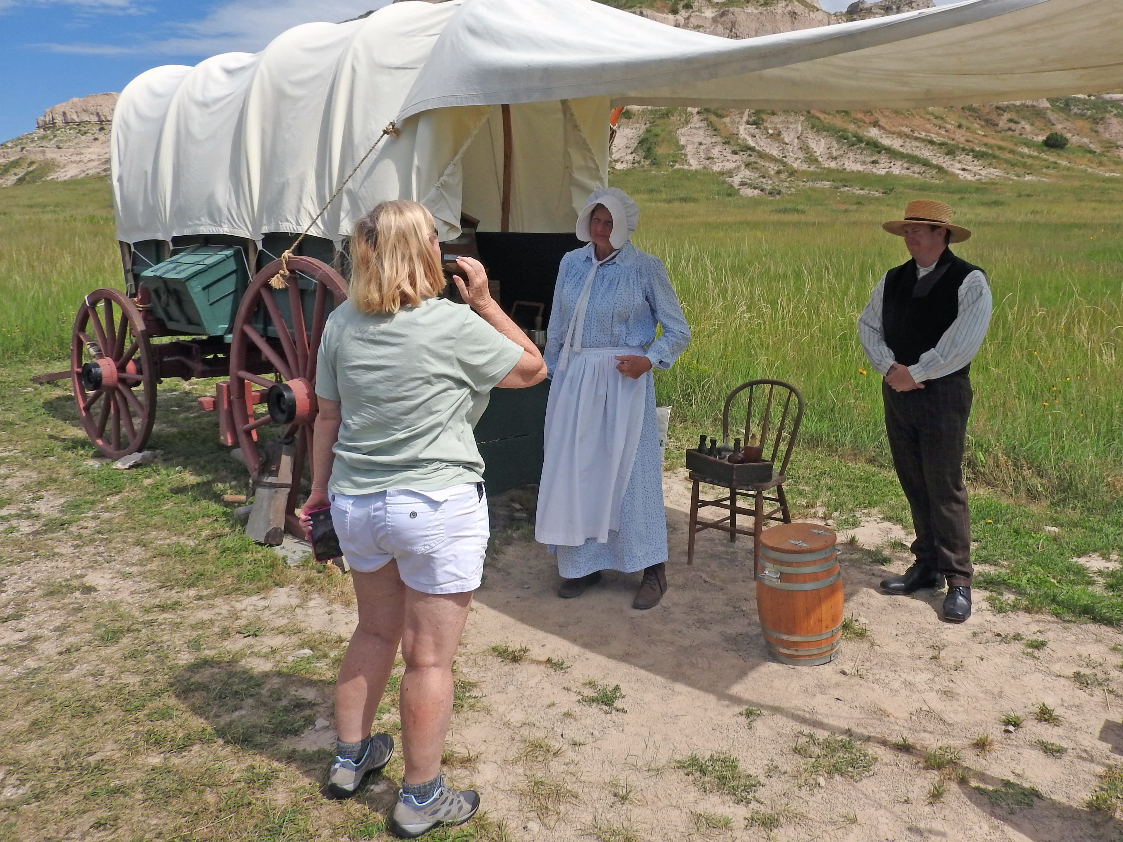 Two rangers, dressed as emigrants on the Oregon Trail, talk with a visitor next to a covered wagon.