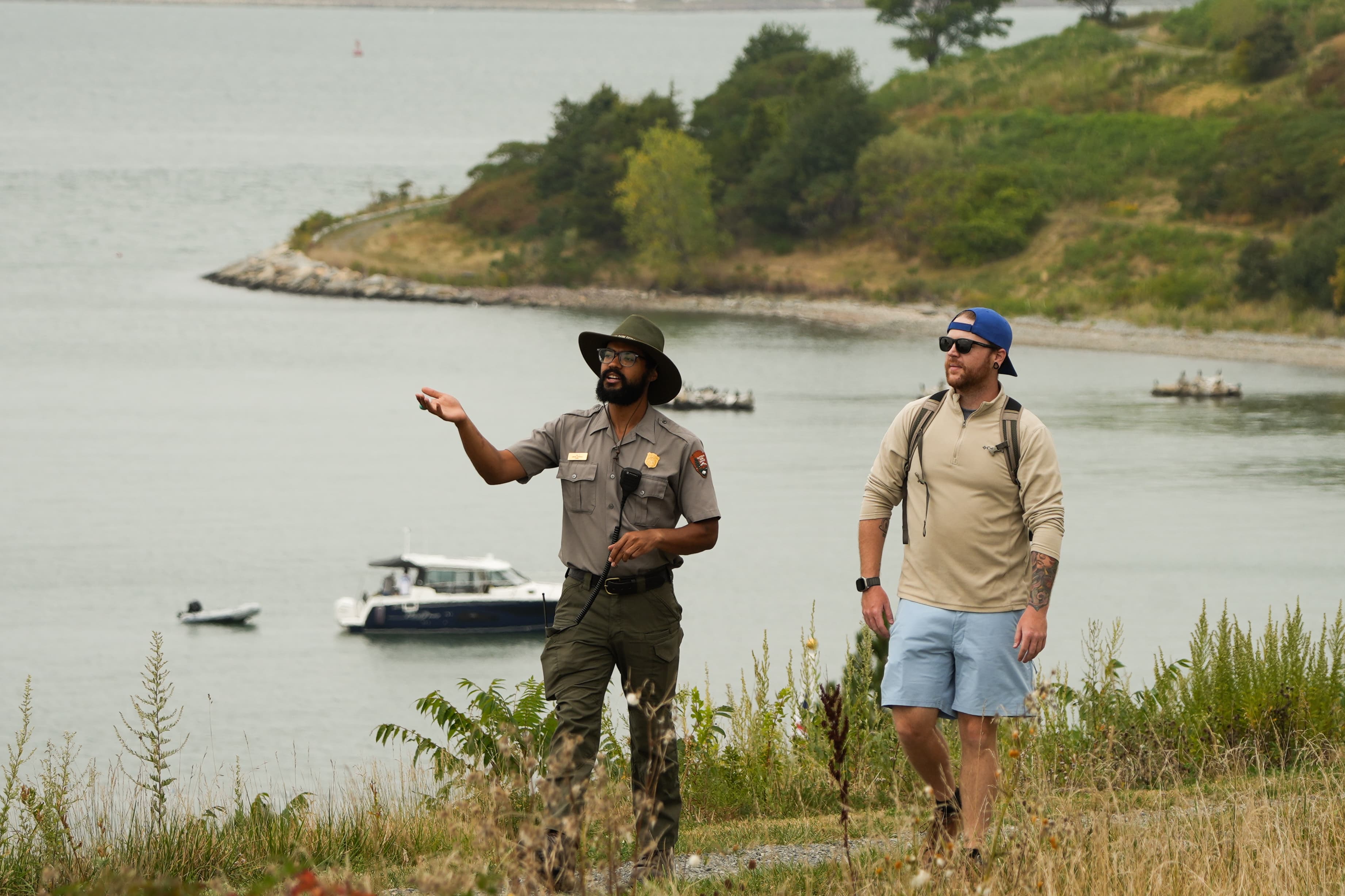 A uniformed ranger walks with a guest on a grassy path on and island, boats in the water behind them