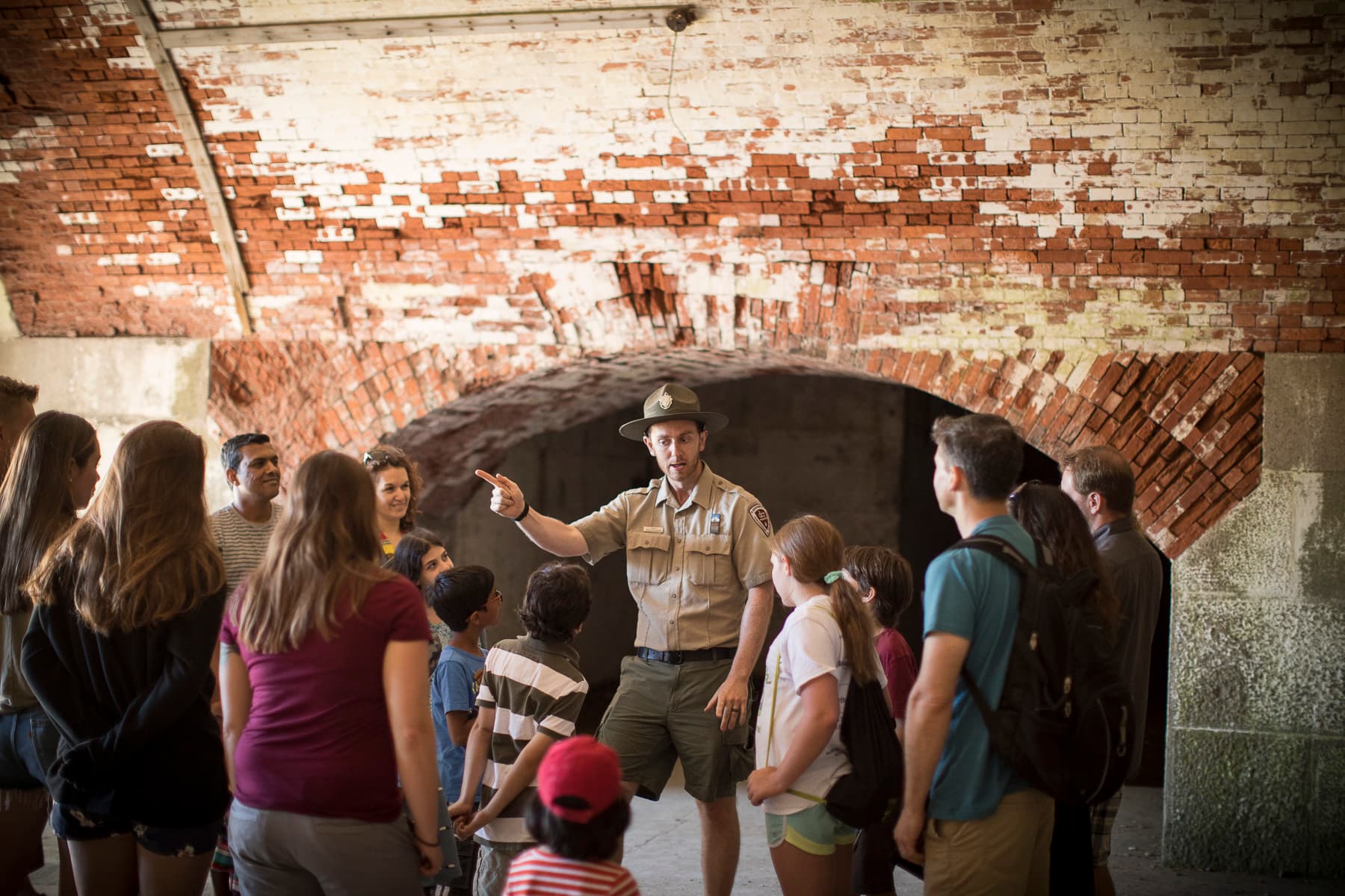 A uniformed ranger speaks to a crowd of mixed ages in an old brick hallway.