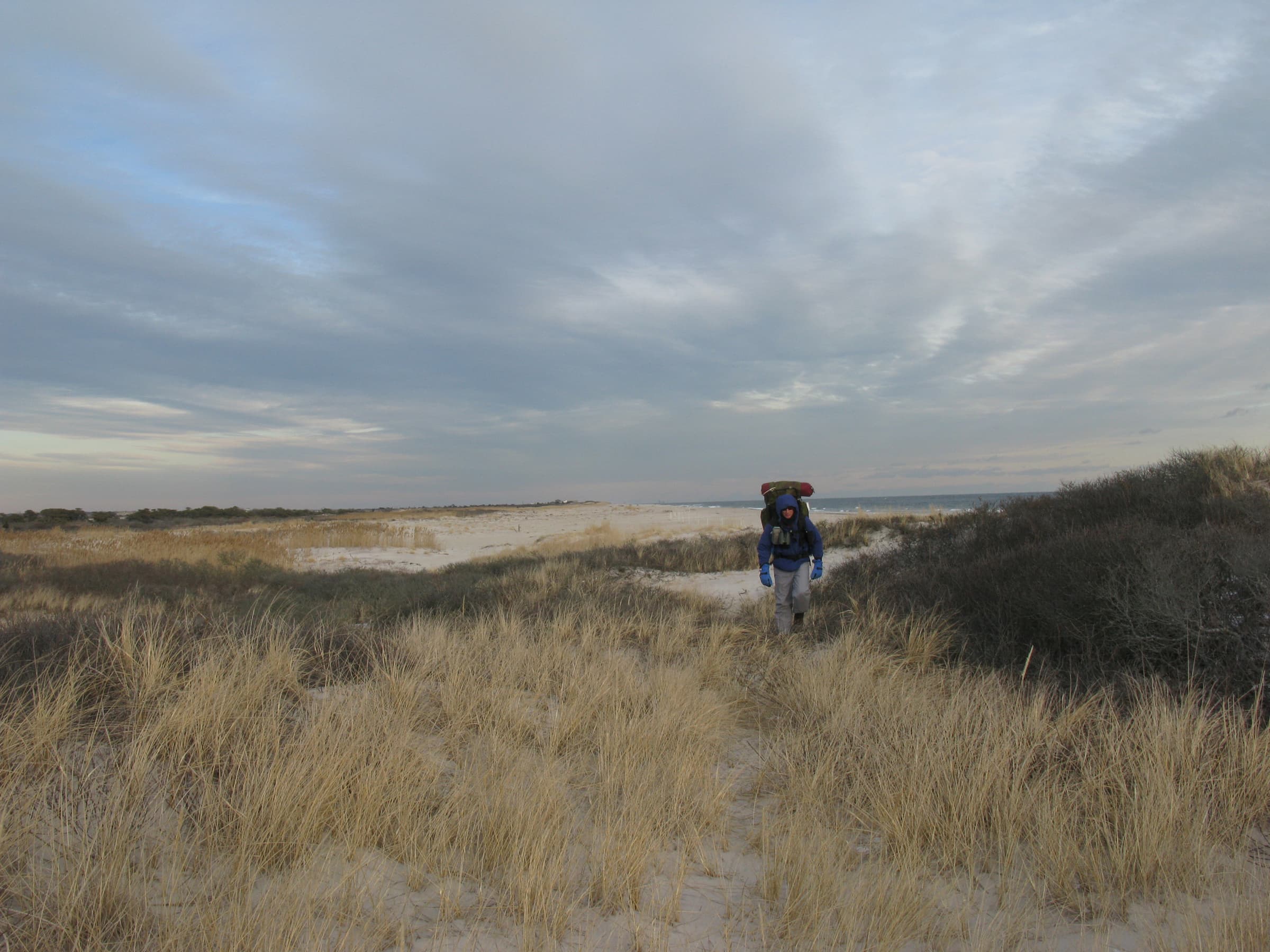 A man with a large backpack walks through sandy trails, dunes, and thickets.