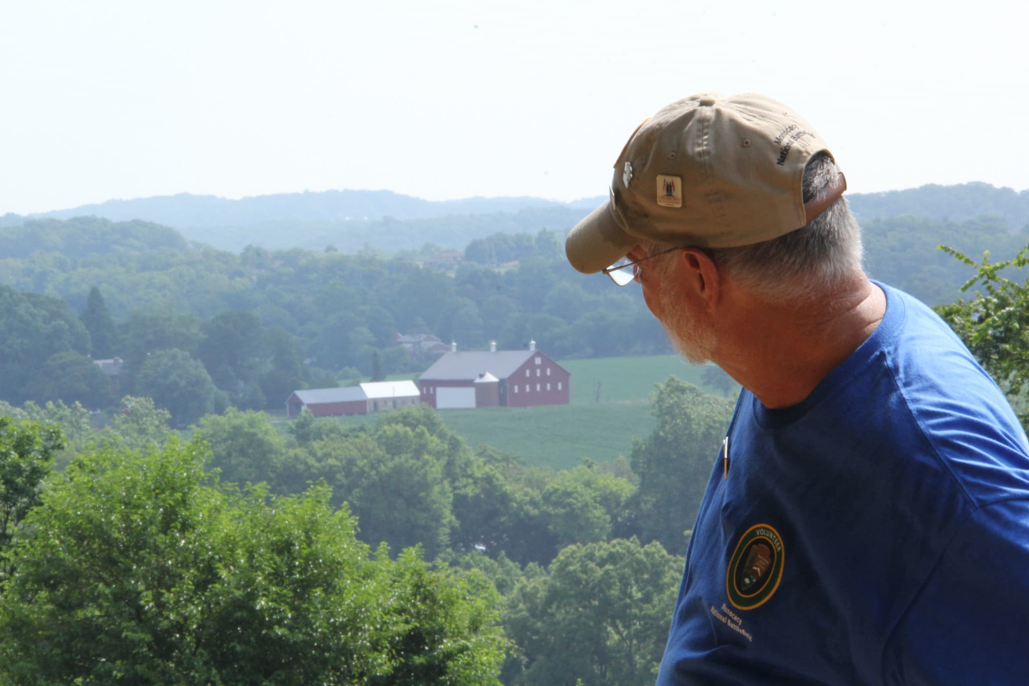 A white male faces away from camera and looks at a red barn and farm buildings in distance below.