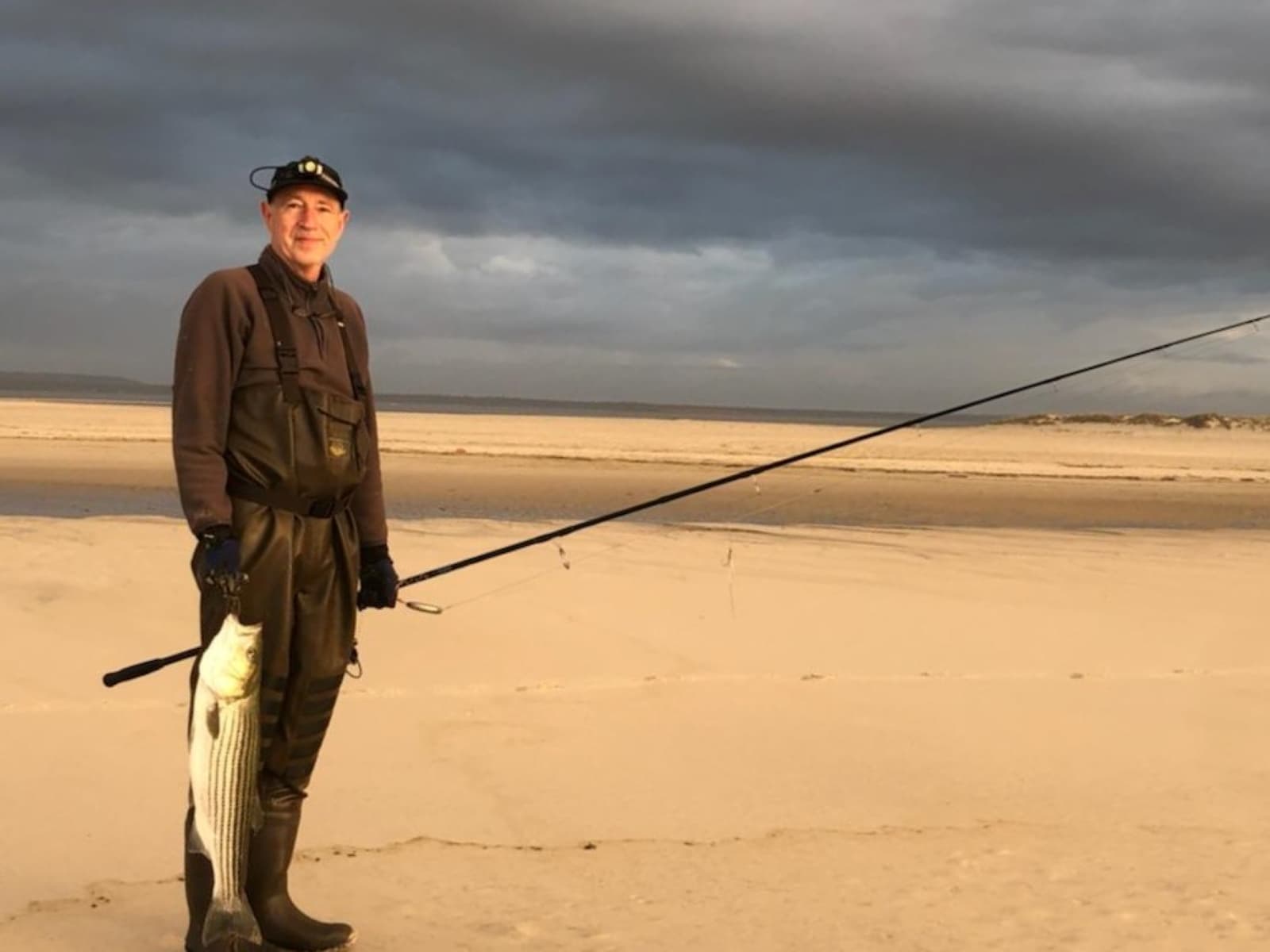 A fisherman stands smiling on an ocean beach with a large stripped bass he caught.