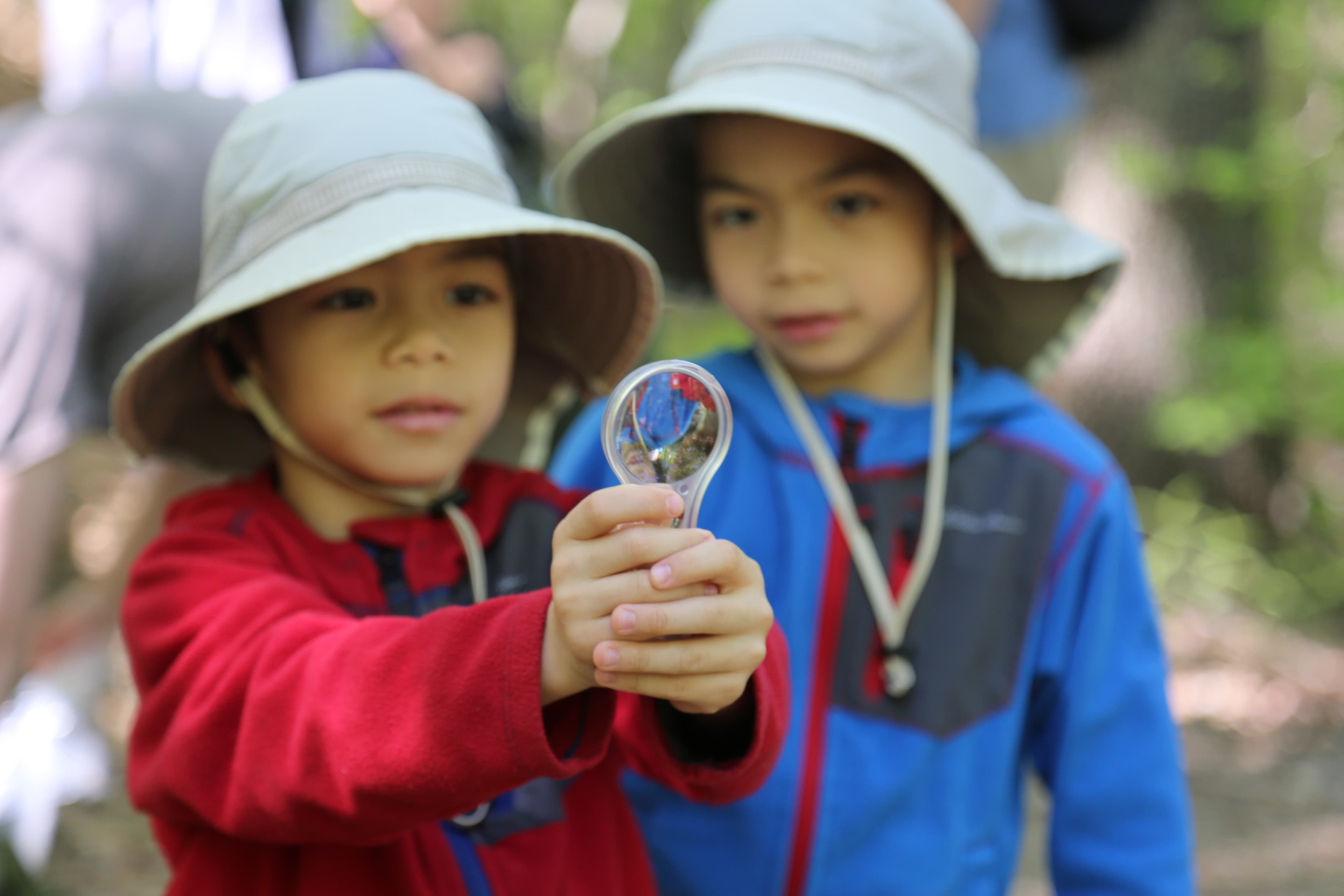 Two children in long sleeved shirts and hats hold up a magnifying lens