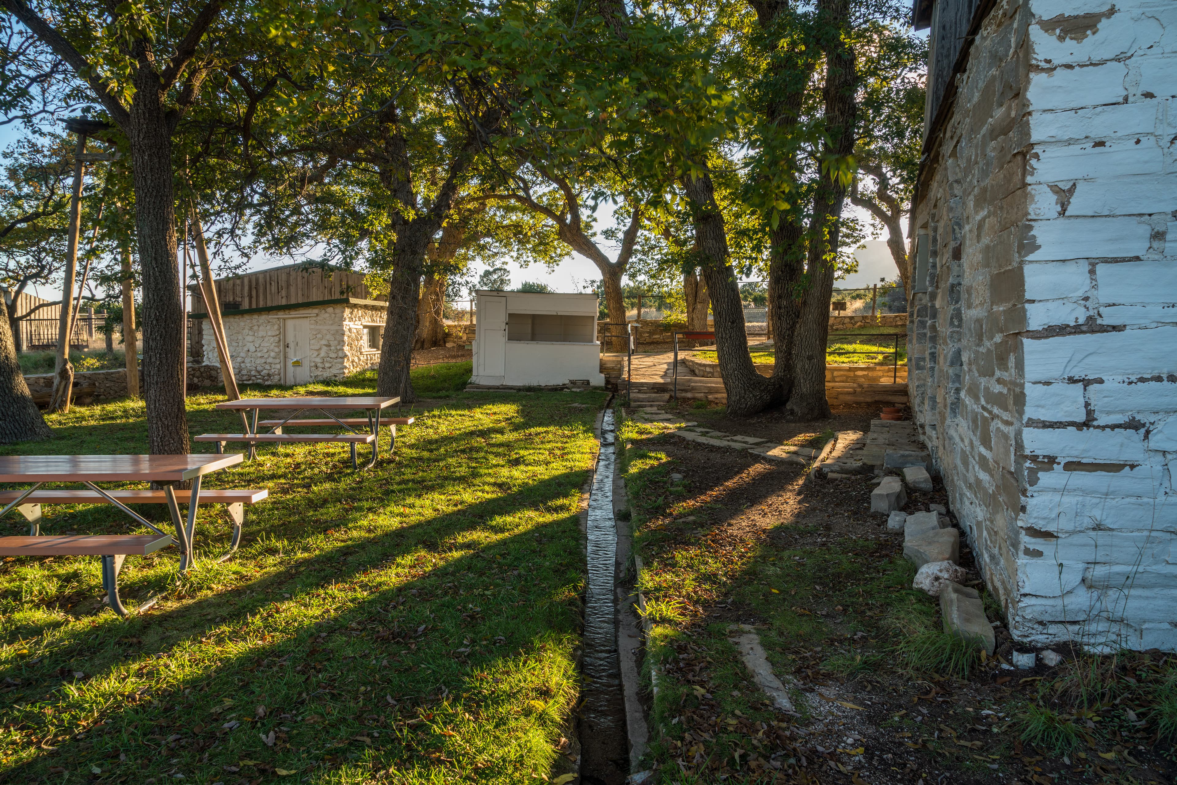 The setting sun casts light and shadows over buildings and a fenced yard with trees.