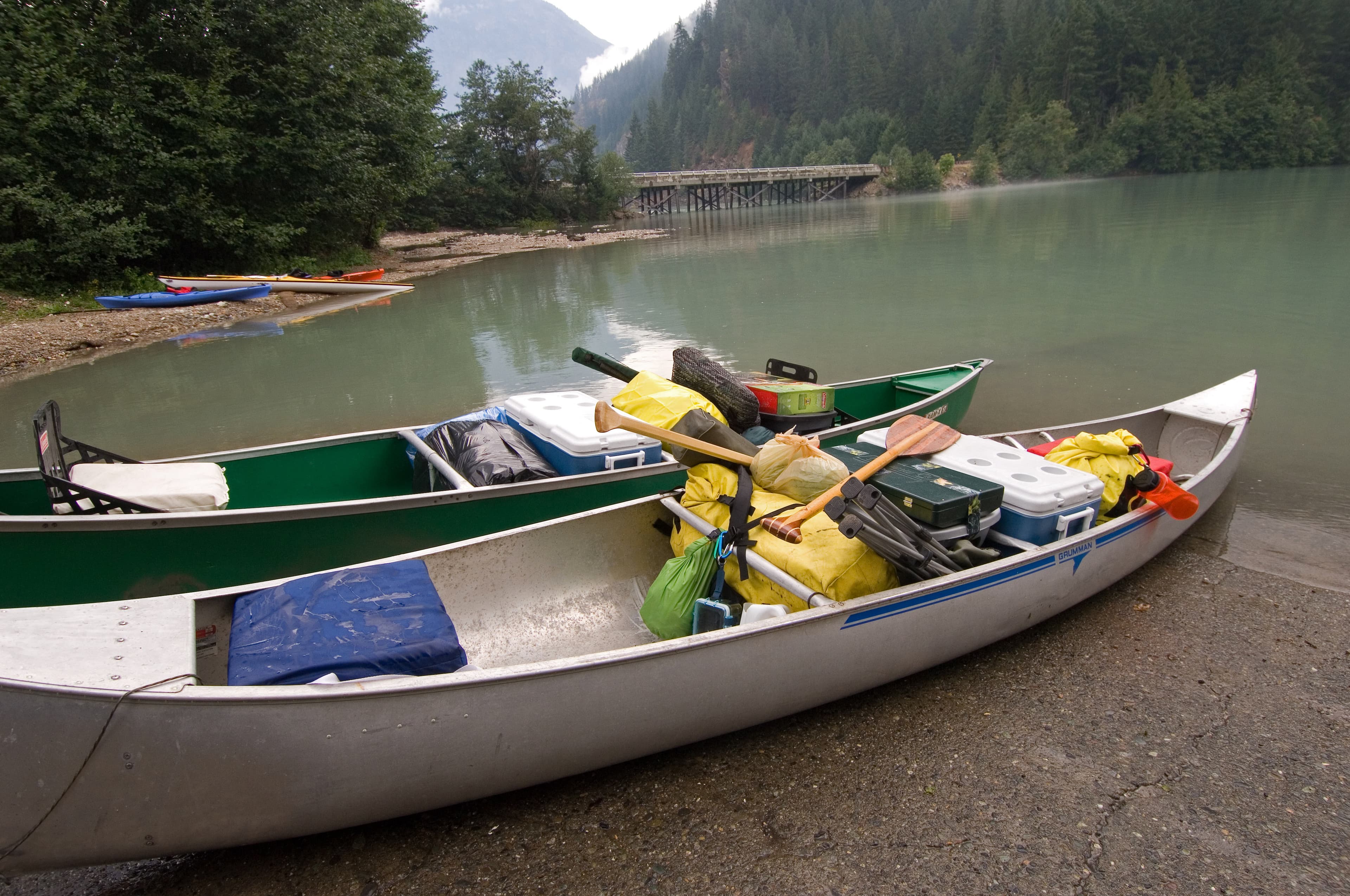 Two silver canoes loaded with camping gear sit on the shore of a lake.