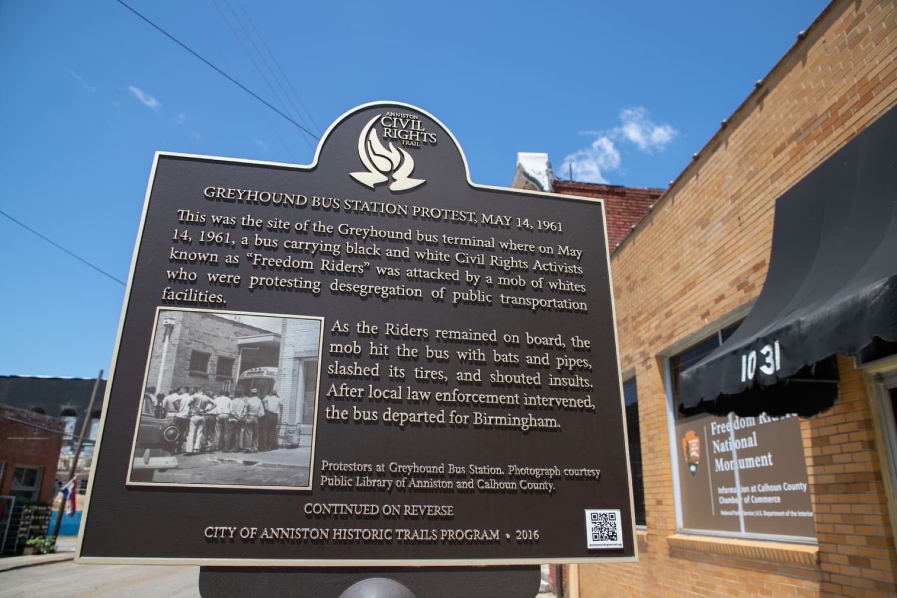 A brown and gold historic marker. In the background: a yellow-brick building with a black awning.
