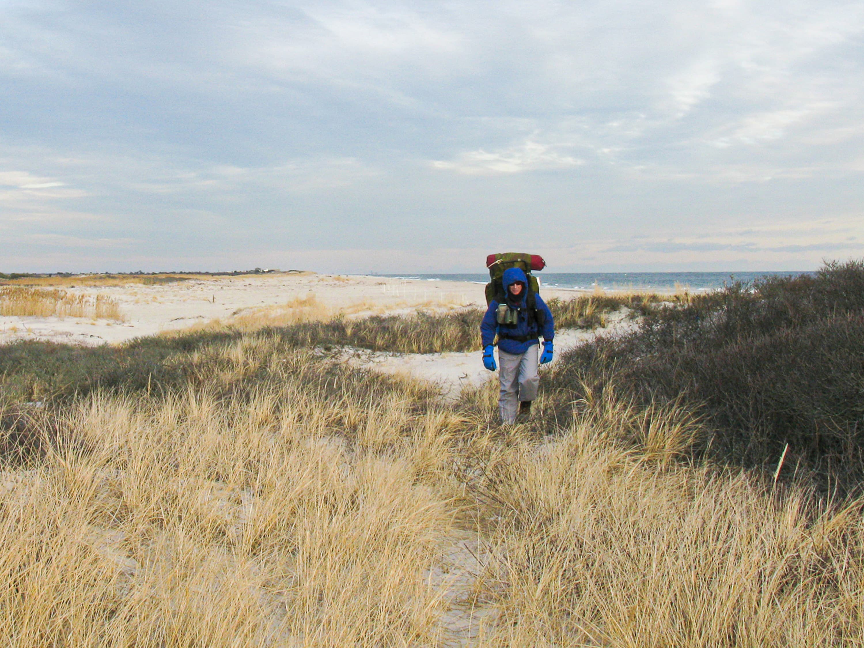 A backpacker with winter clothes walks a grassy dune near the ocean.