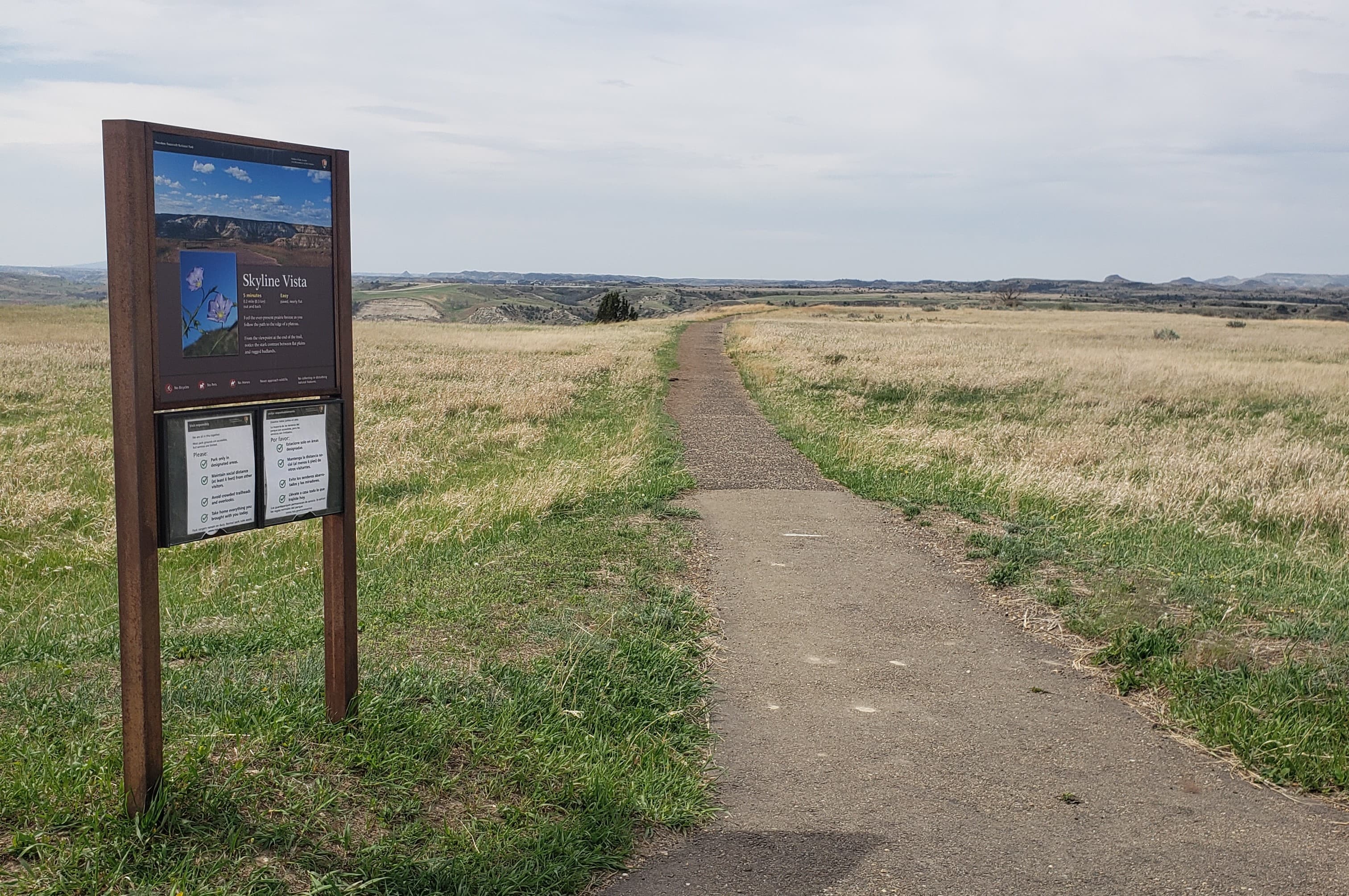A trailhead sign next to a paved trail in a grassy area. Buttes are visible on the horizon.