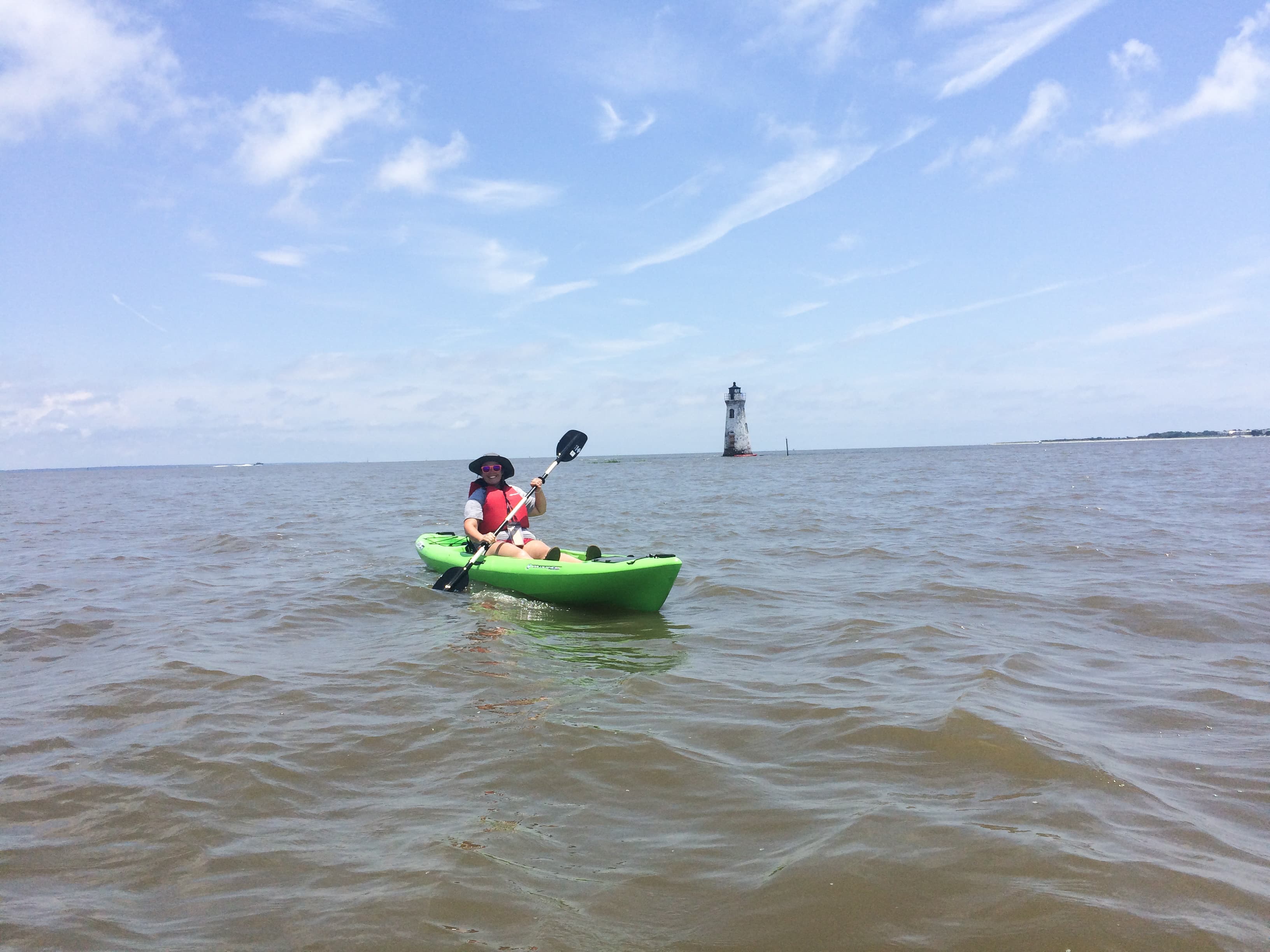 A woman in a green kayak with a lighthouse distantly behind.