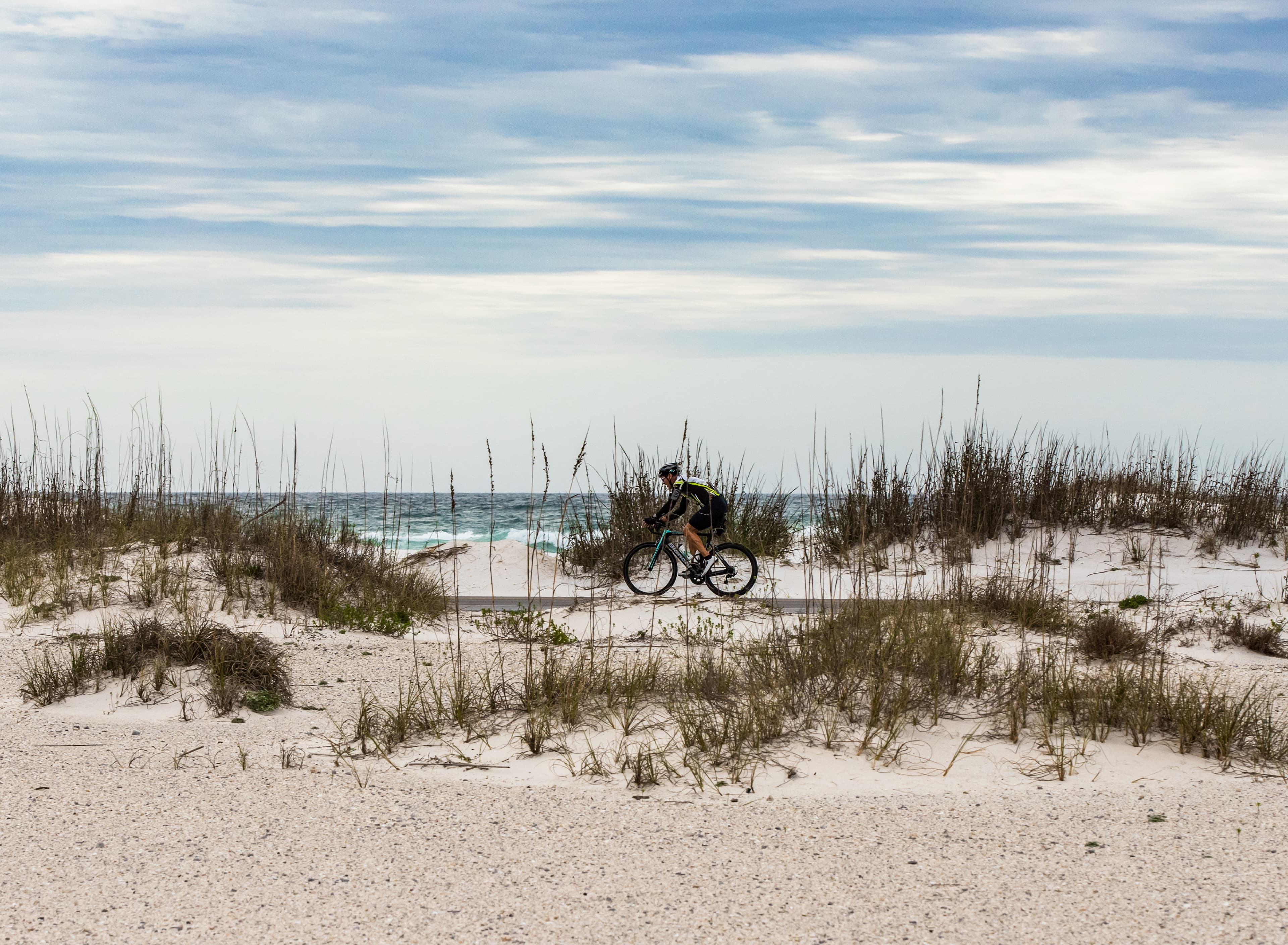 A bicyclist pedals on the road with sand dunes in the foreground and the ocean behind.