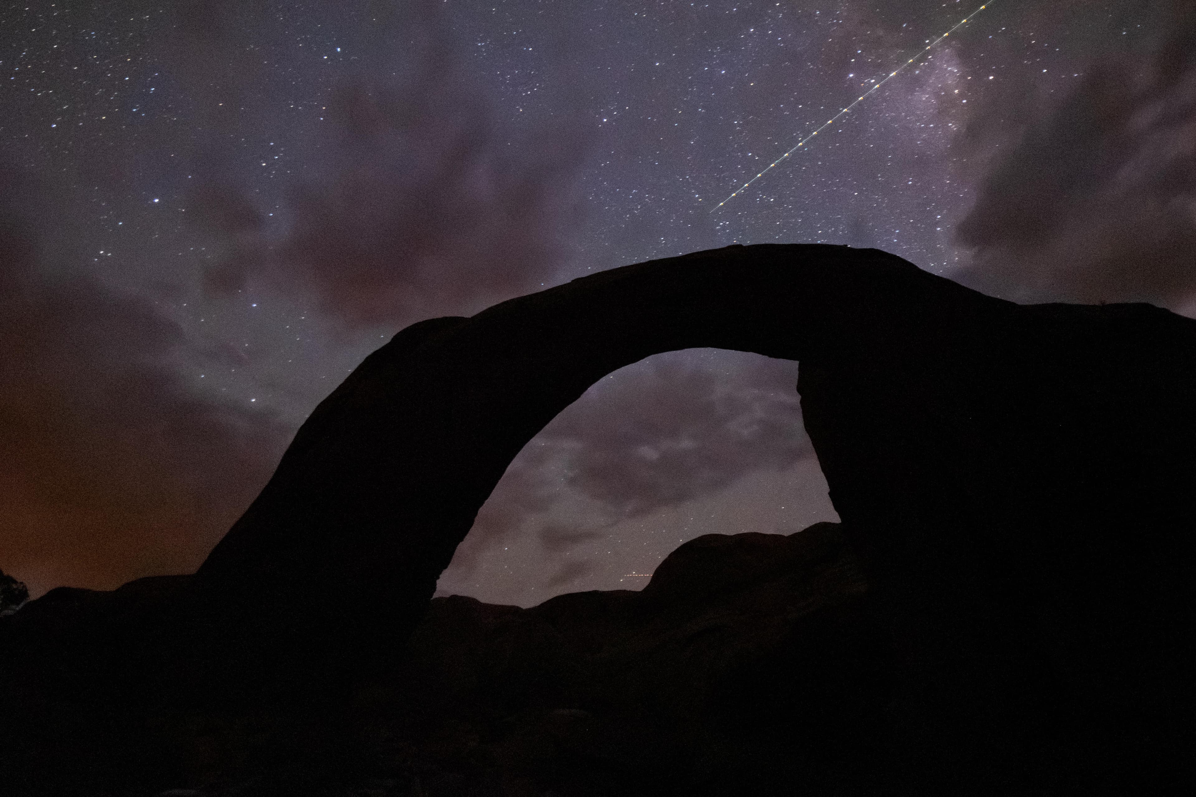 A silhouetted rock arch under a stary sky with a satellite flying by.