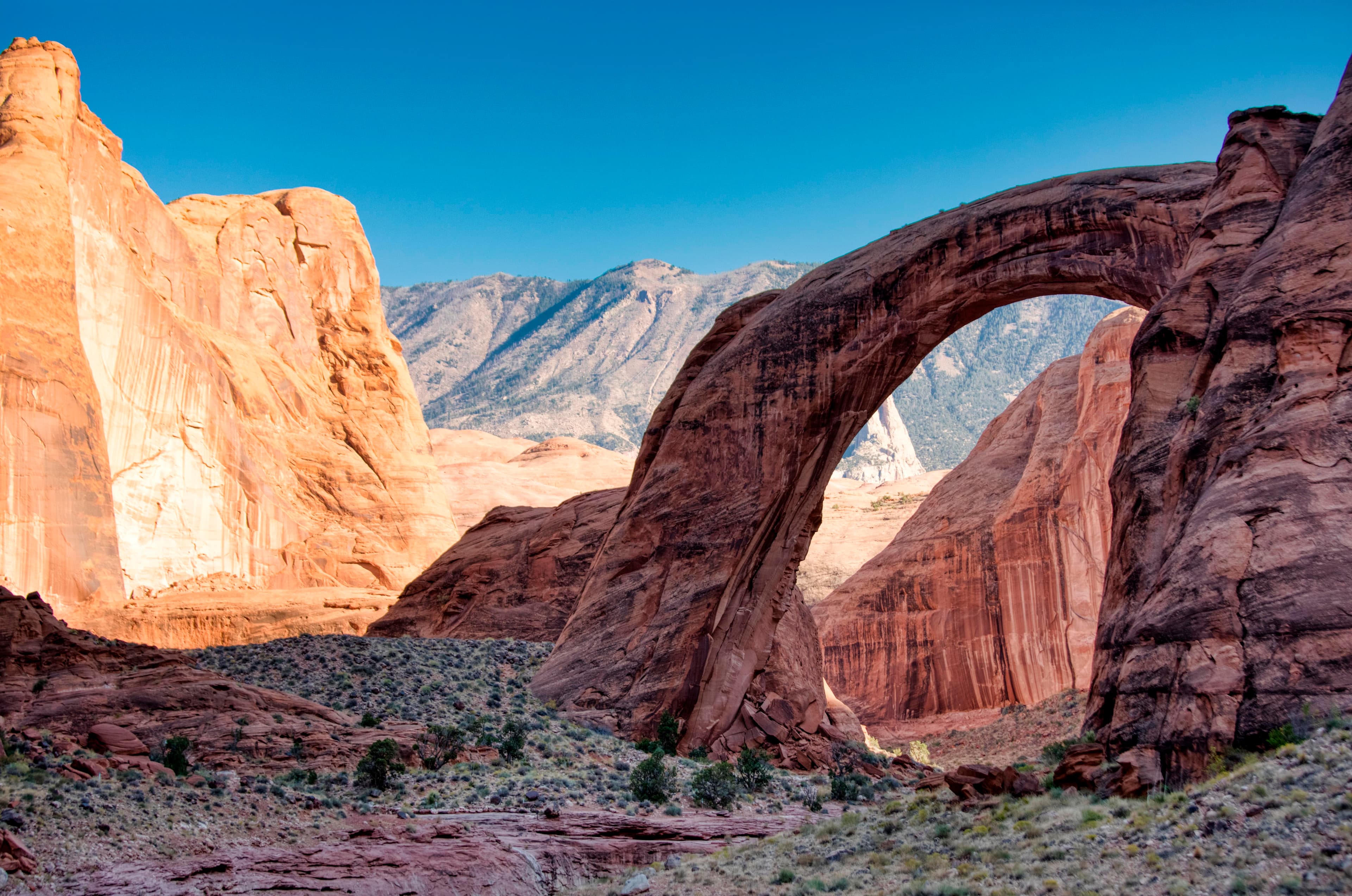 A natural stone arch sits between red rock canyon walls. A mountain is seen through the arch.