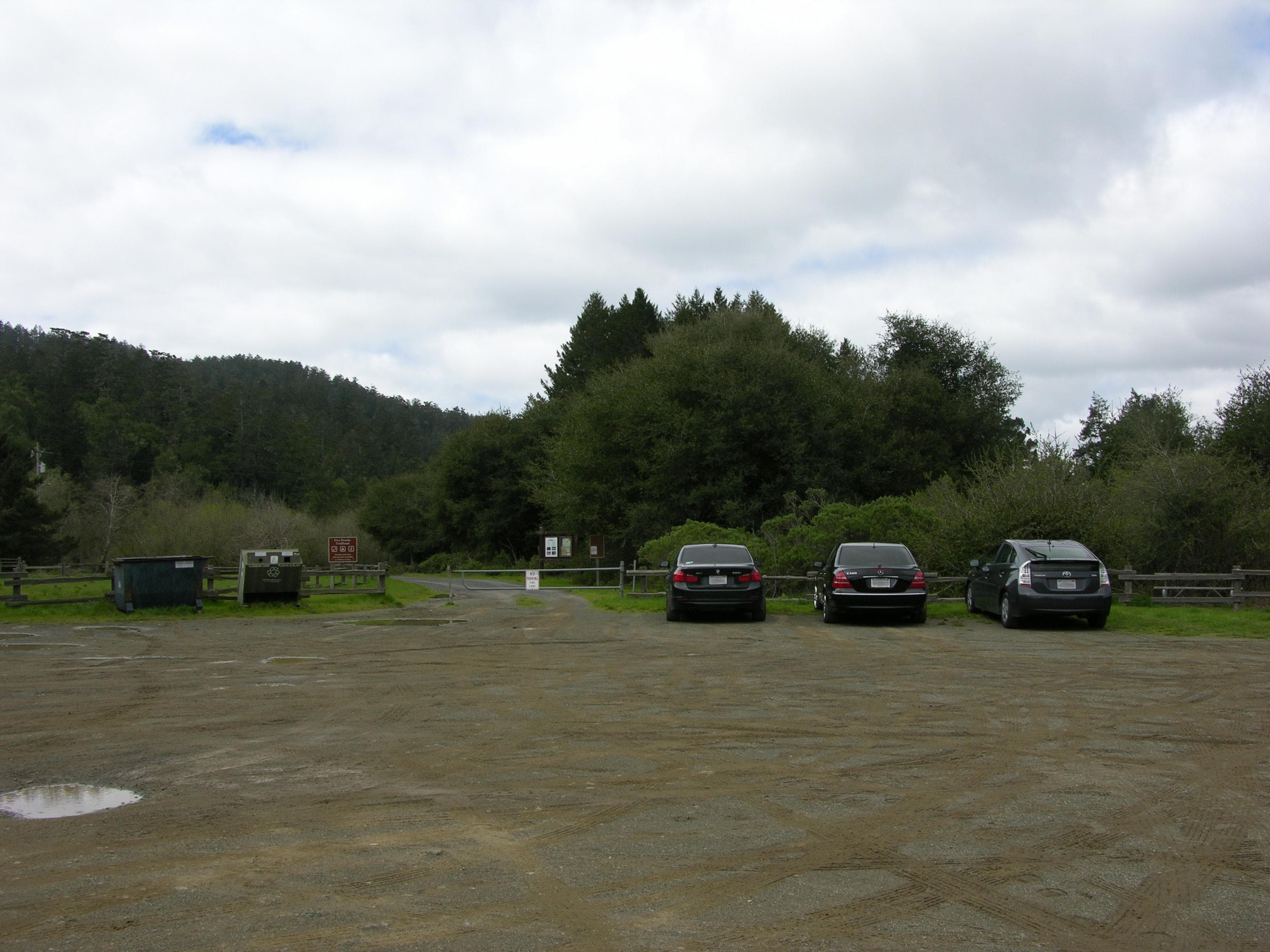 Three vehicles parked in a dirt lot. On the left is a dirt road blocked by a gate.