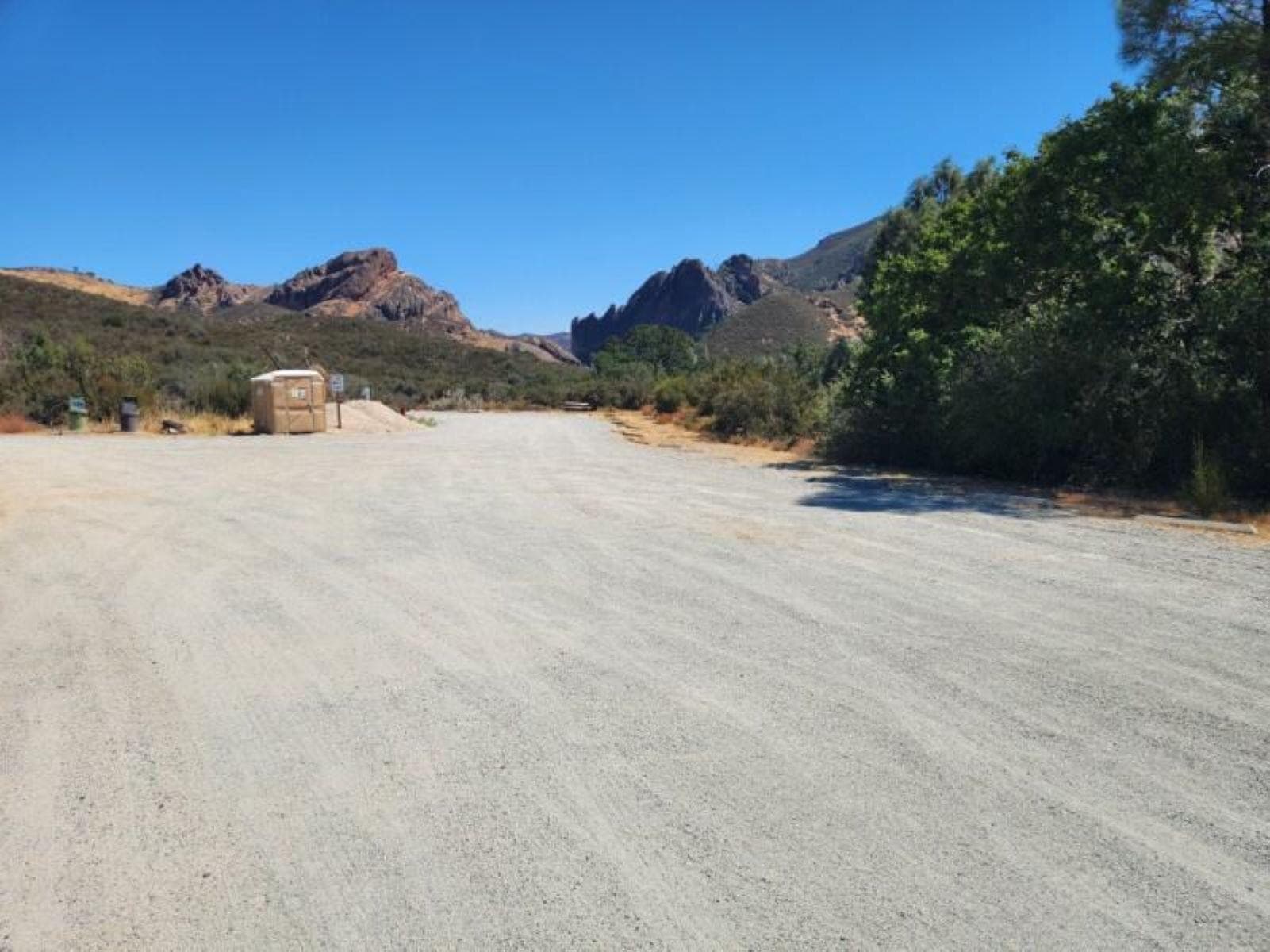 Rock formations seen behind thick chaparral covered hillsides from a gravel parking area.