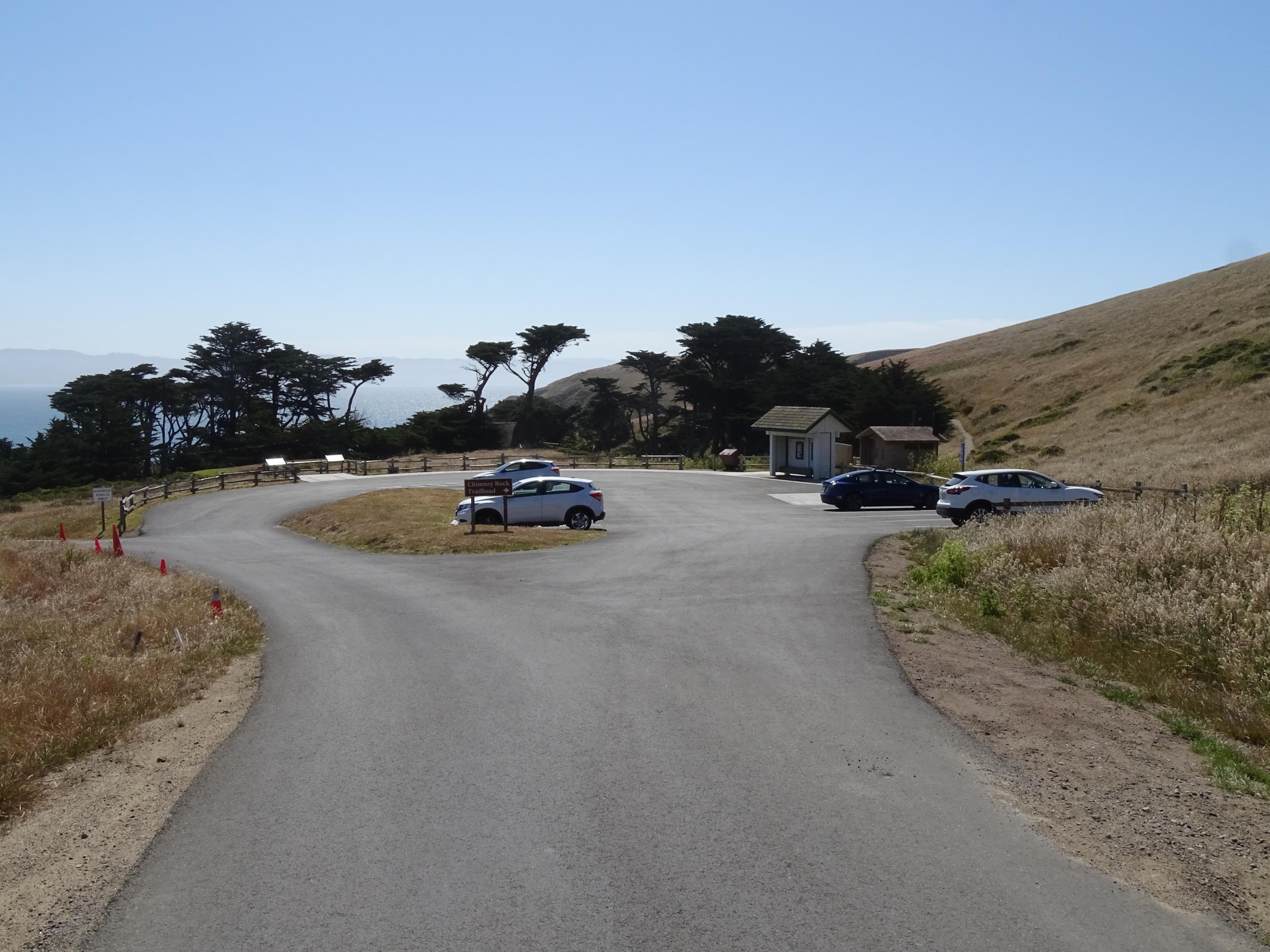Four vehicles in a small paved parking lot adjacent to a small white shelter and a vault toilet.