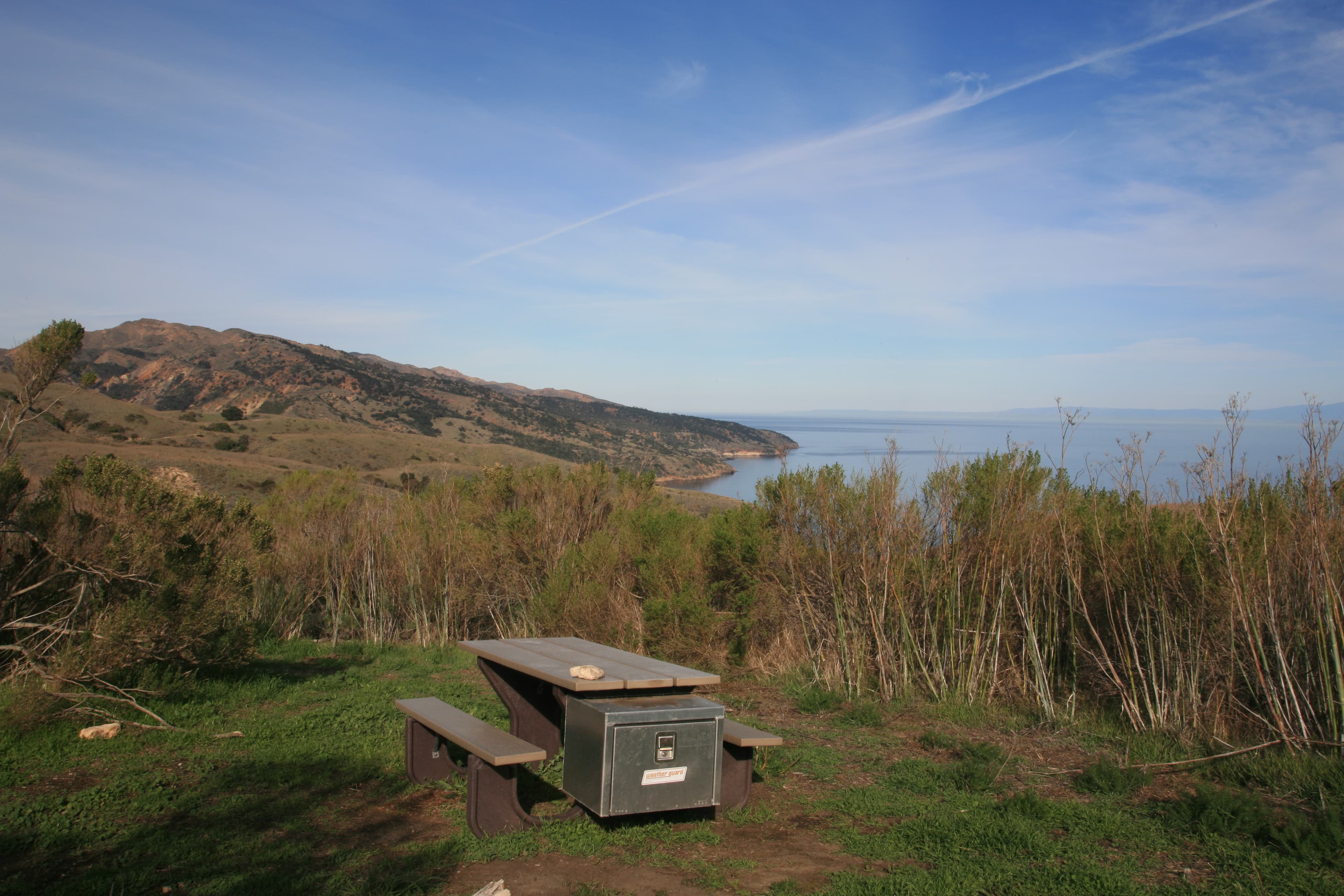 Campground picnic table on green grass overlooking ocean, coastline, and blue sky with white clouds