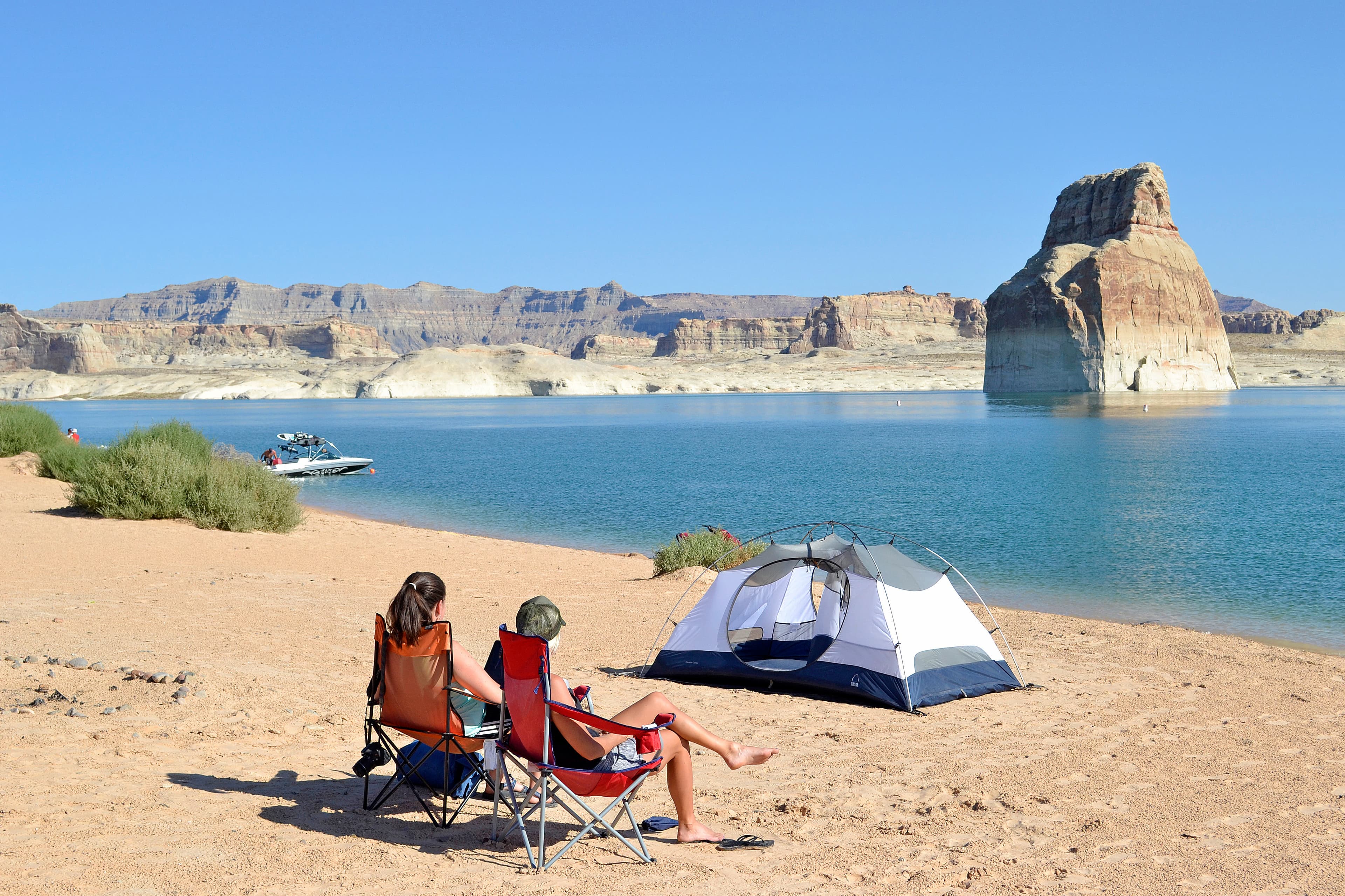 Two people sit in beach chairs in front of a tent pitched on a beach near the water's edge.