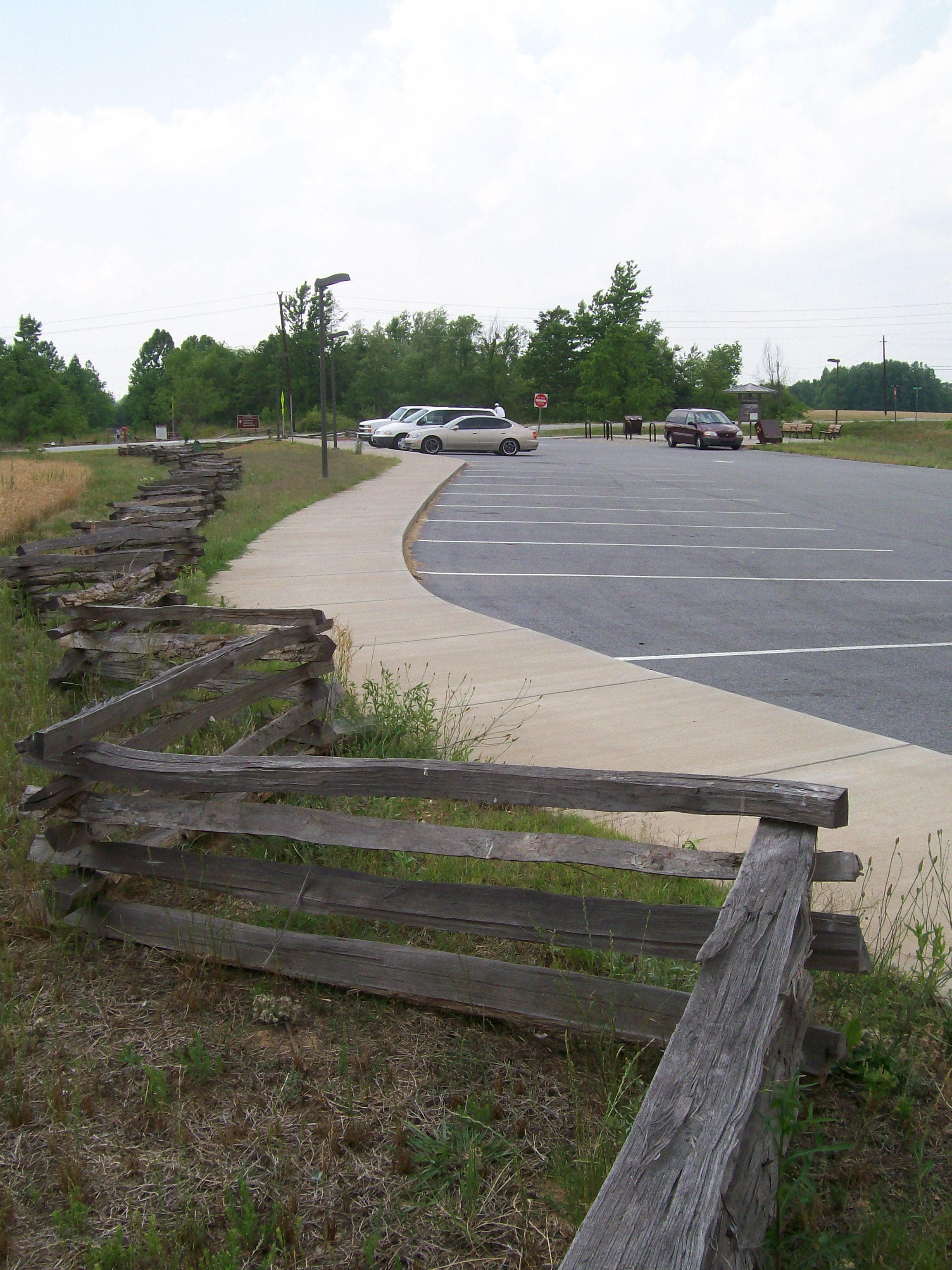 A splitrail fence and sidewalk are to the left of a parking lot.